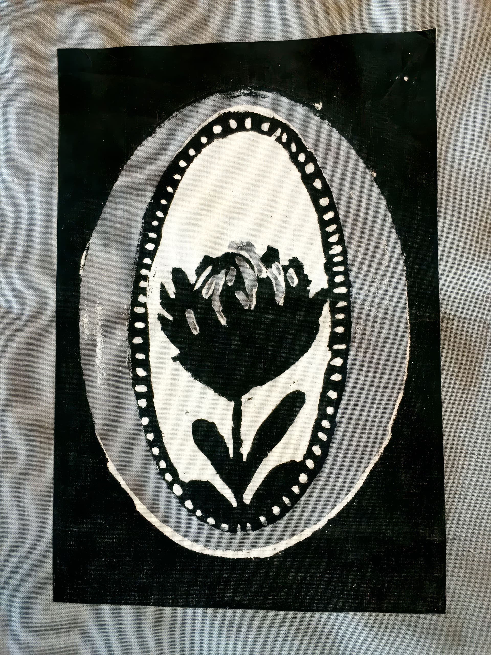 A black and white print depicts a stylised flower with two leaves inside an oval border. The border is decorated with small dots. The flower and borders are surrounded by a solid black rectangular background, set against grey fabric.