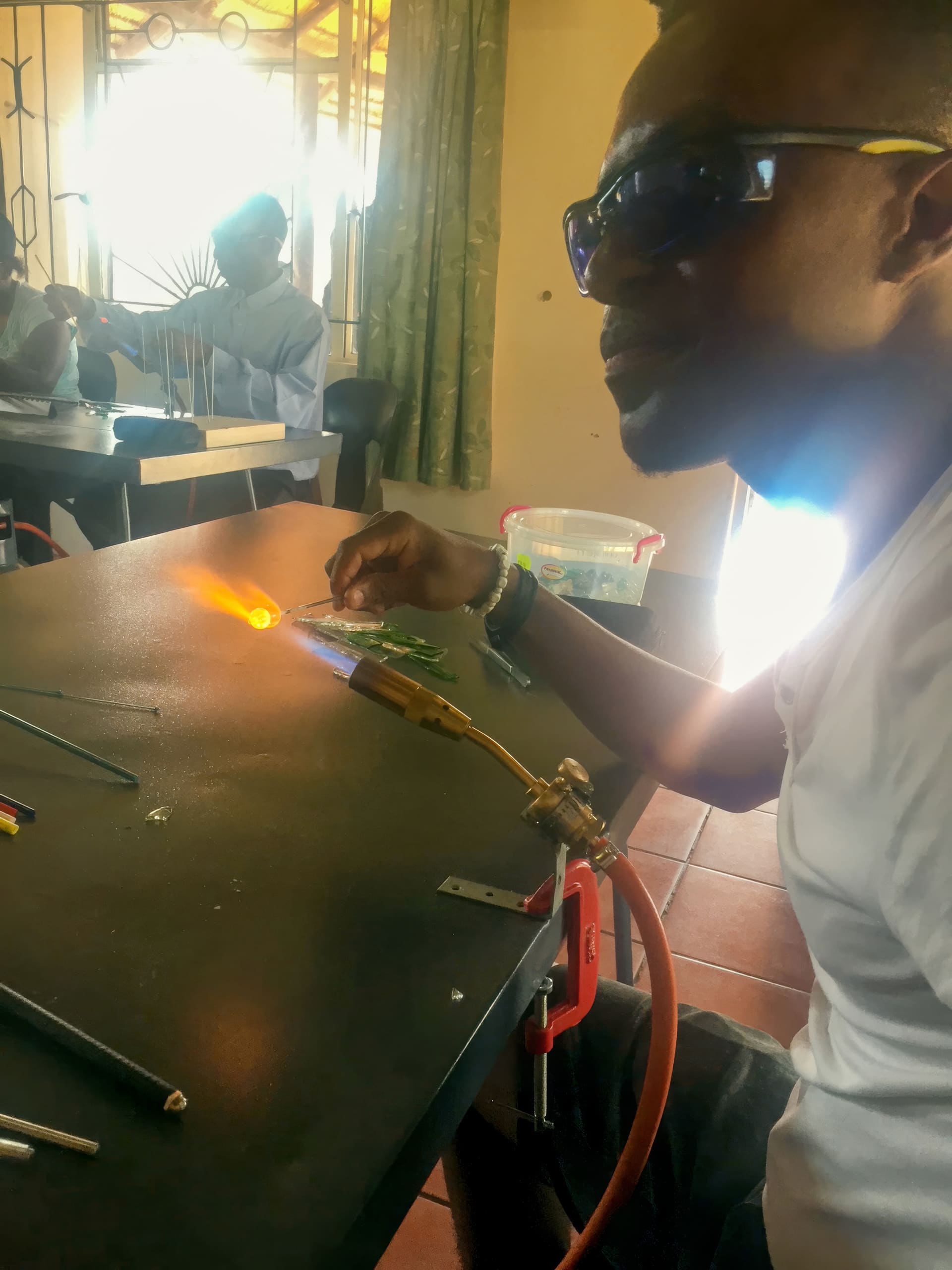 A man wearing dark safety glasses uses a blowtorch to heat a rod on a black table in a sunlit room. Other people work at tables in the background. Tools and coloured rods are scattered on the table near him.