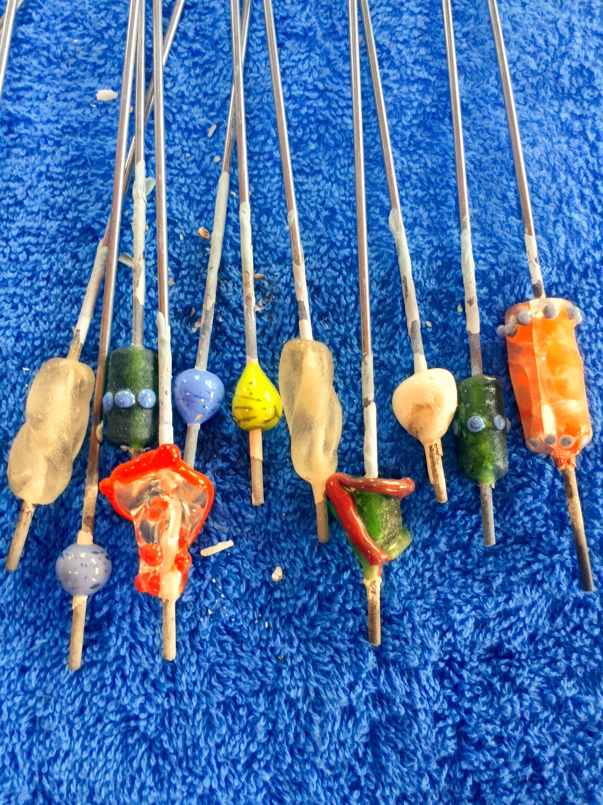 Several metal rods with colourful, hand-shaped glass beads are arranged on a bright blue textured towel. The beads are various shapes and colours, including green, orange, yellow, blue, and beige, some with dots and layered designs.