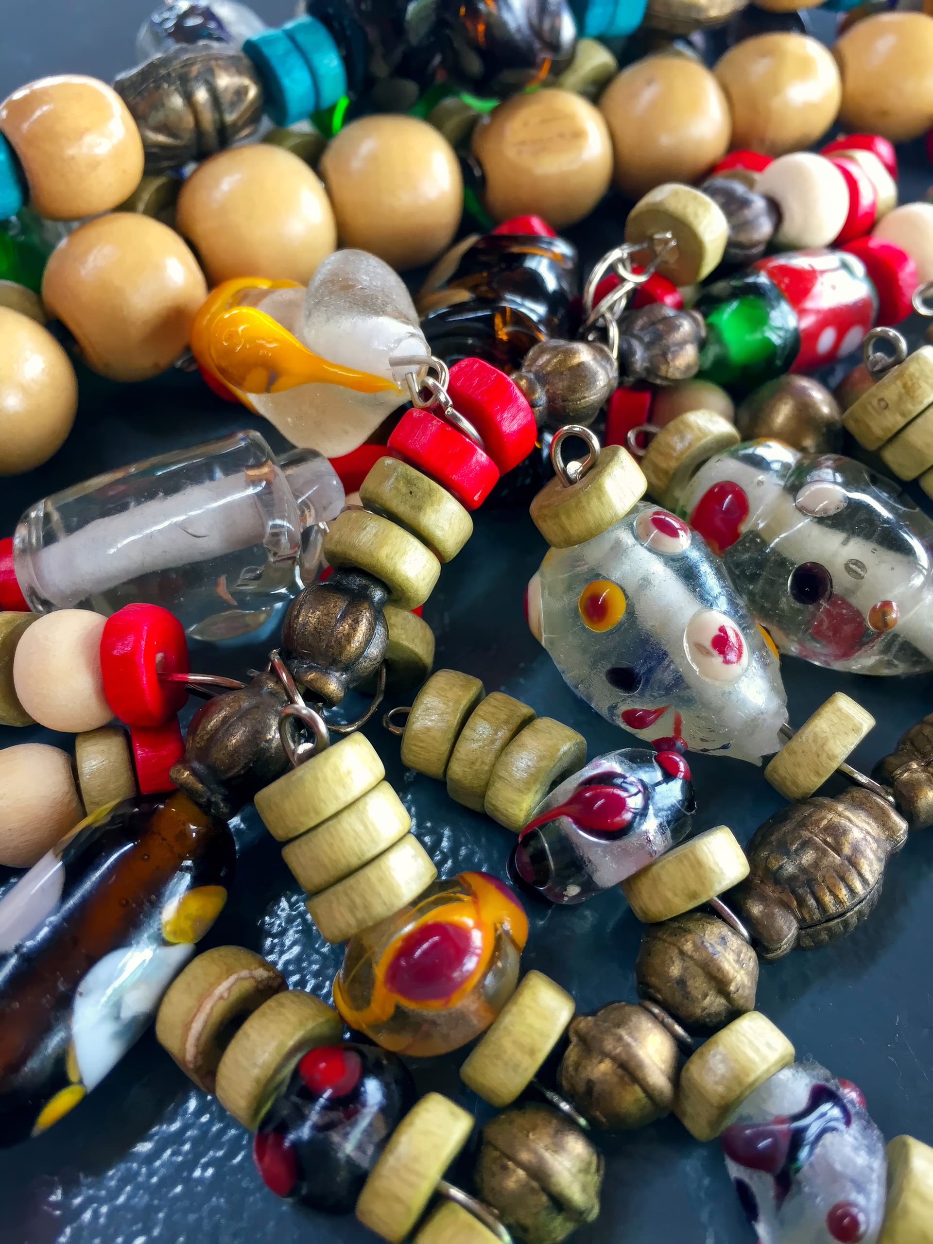 A close-up of colourful beaded necklaces featuring round wooden, metallic, and glass beads, including clear glass beads with red and yellow patterns, green and red beads, and some with heart and capsule shapes, all arranged in a tangled pile.