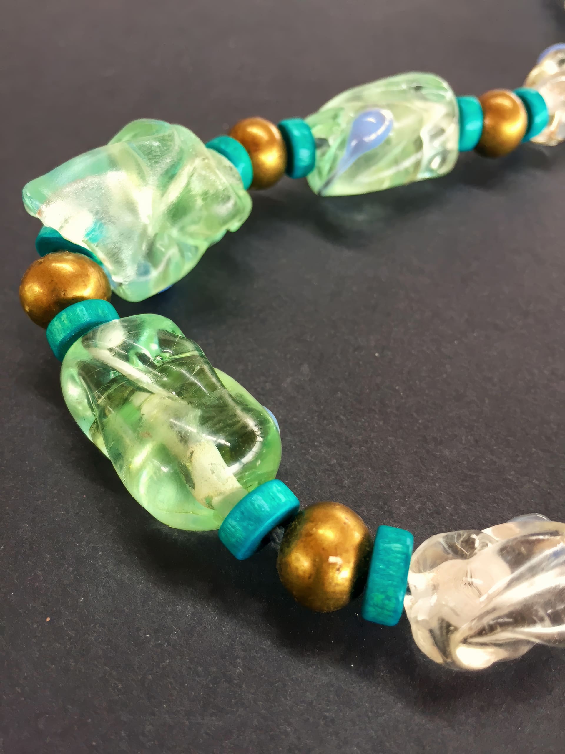 Close-up of a beaded necklace featuring large, irregularly shaped translucent green and clear beads, metallic gold round beads, and small flat turquoise discs, arranged in a repeating pattern on a black background.