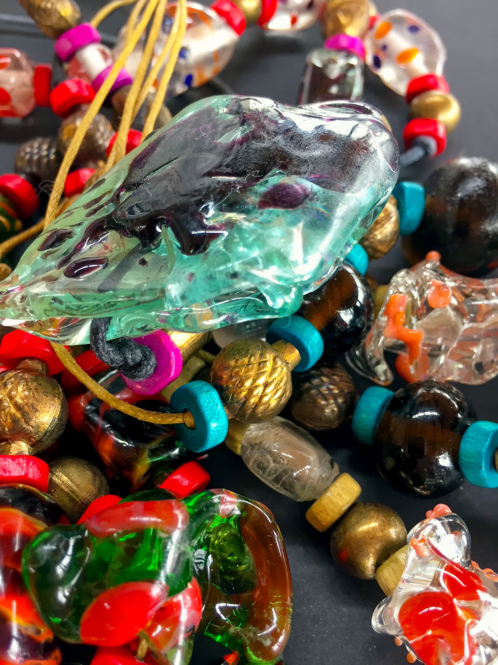 A close-up of colourful glass and wooden bead necklaces, featuring vibrant red, turquoise, and gold beads, and several large glass pendants in green, clear, and multicoloured hues, all set against a dark background.