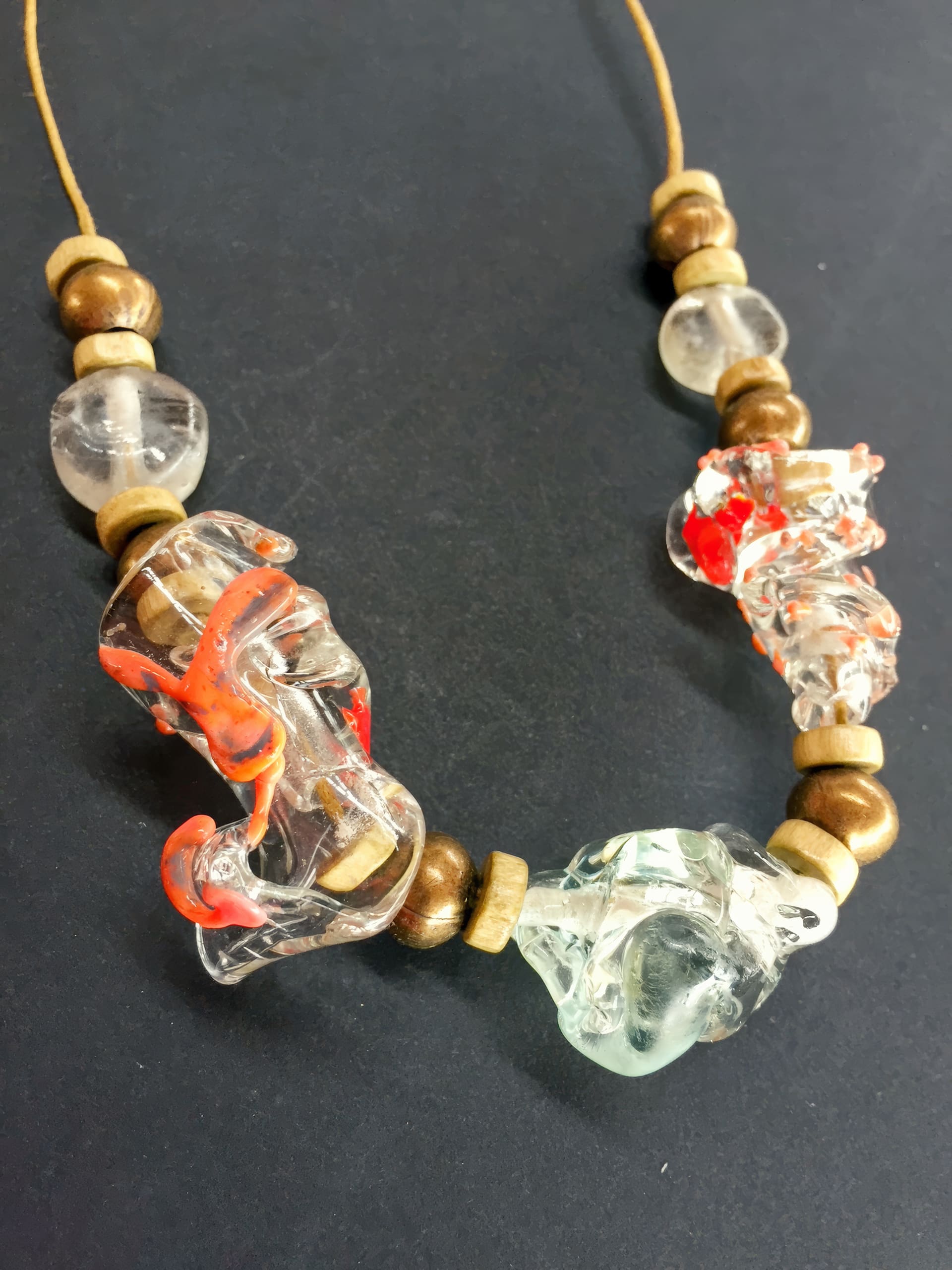 A necklace with chunky, irregular glass beads—some clear, others with red or mint green accents—interspersed with round wooden beads of different sizes, all strung on a tan cord against a black background.