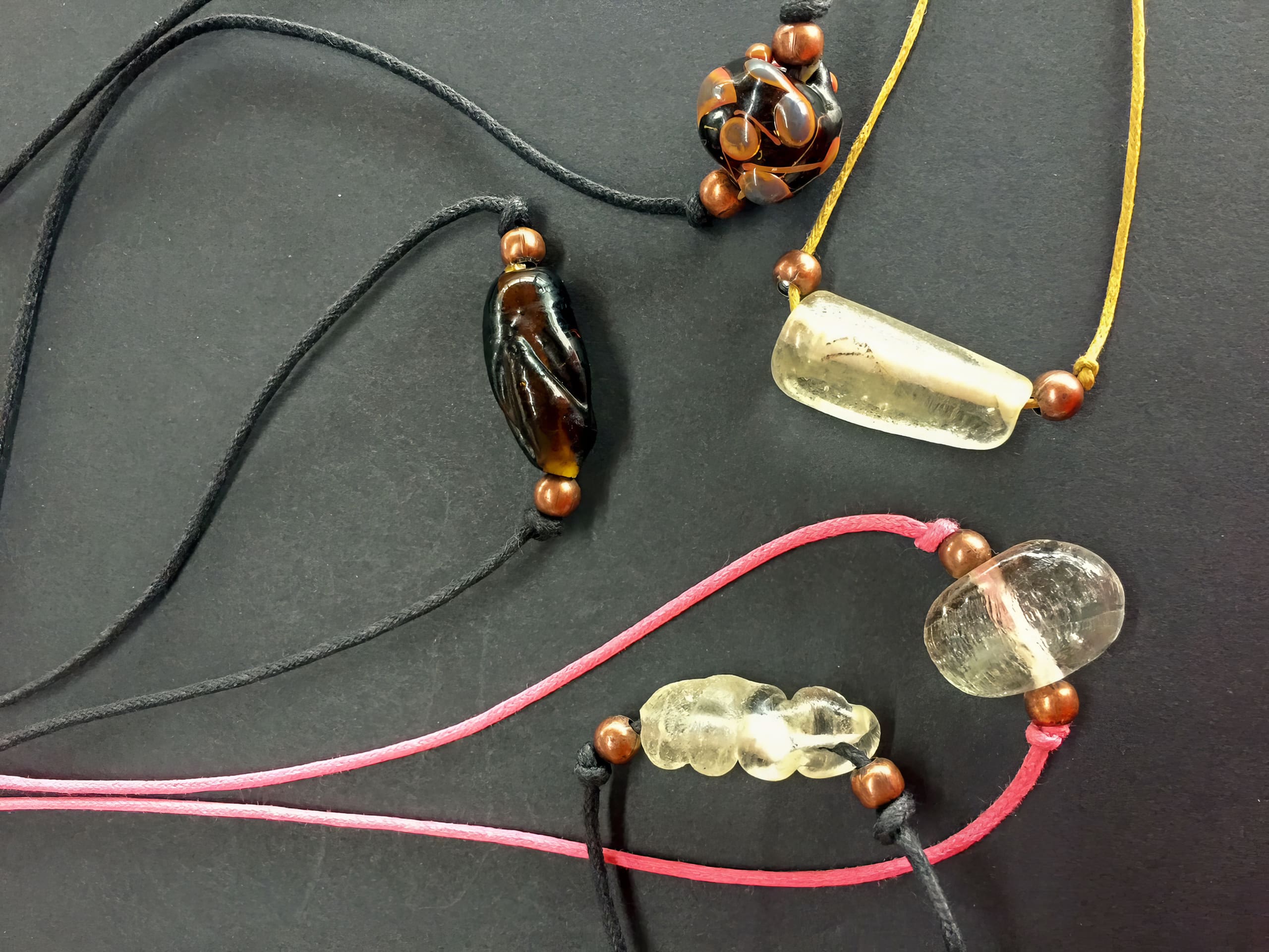 Four handmade necklaces with black, yellow, and pink cords, each featuring a unique polished stone or glass pendant and wooden bead accents, are arranged on a black surface. The pendants vary in colour, including brown, clear, and amber tones.