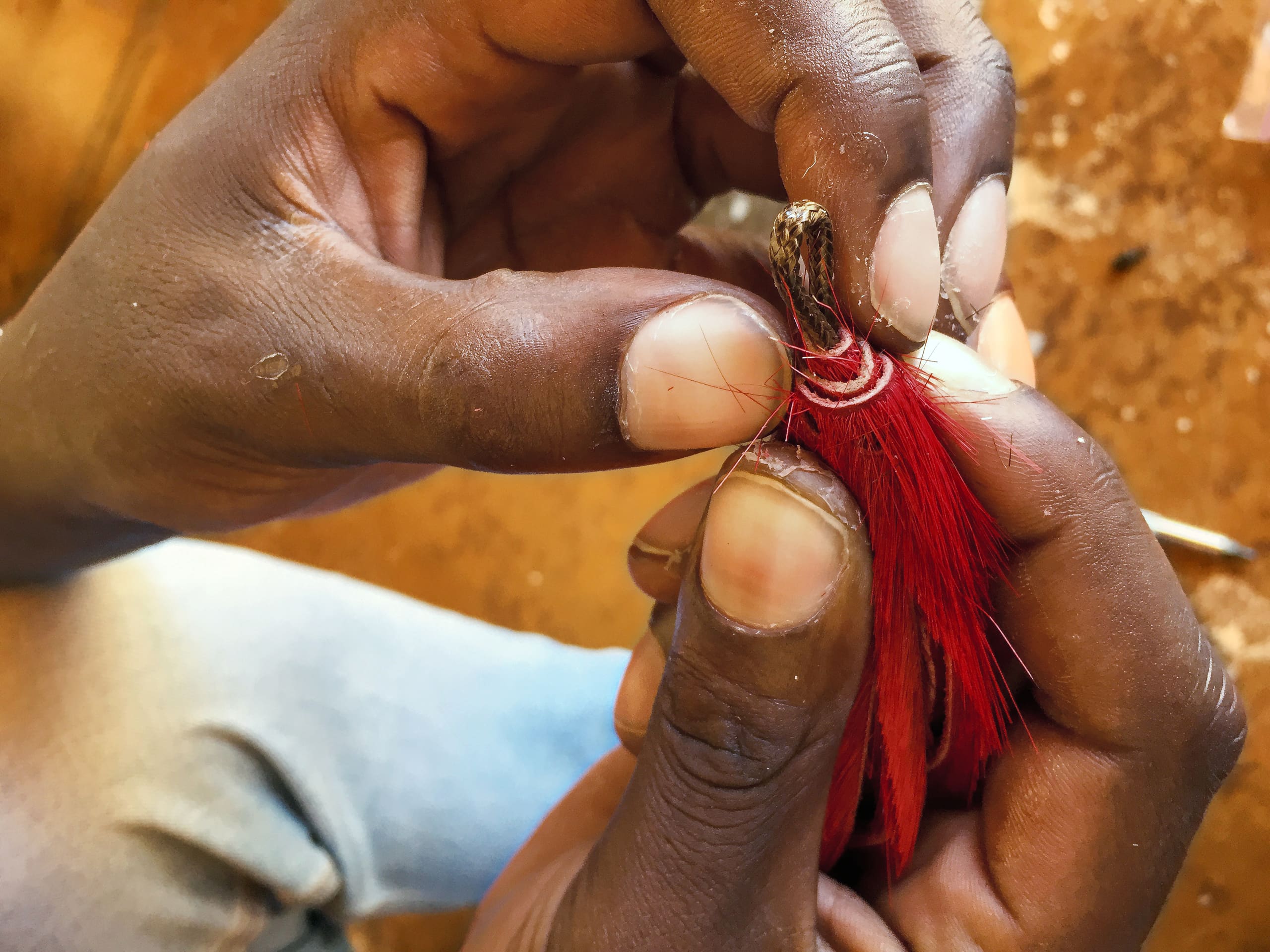 Close-up of two hands carefully tying red feathers to a small object with white string. The background is out of focus, showing brown and tan tones, possibly a wooden surface, and part of a knee in blue jeans. The hands appear skilful and focused.
