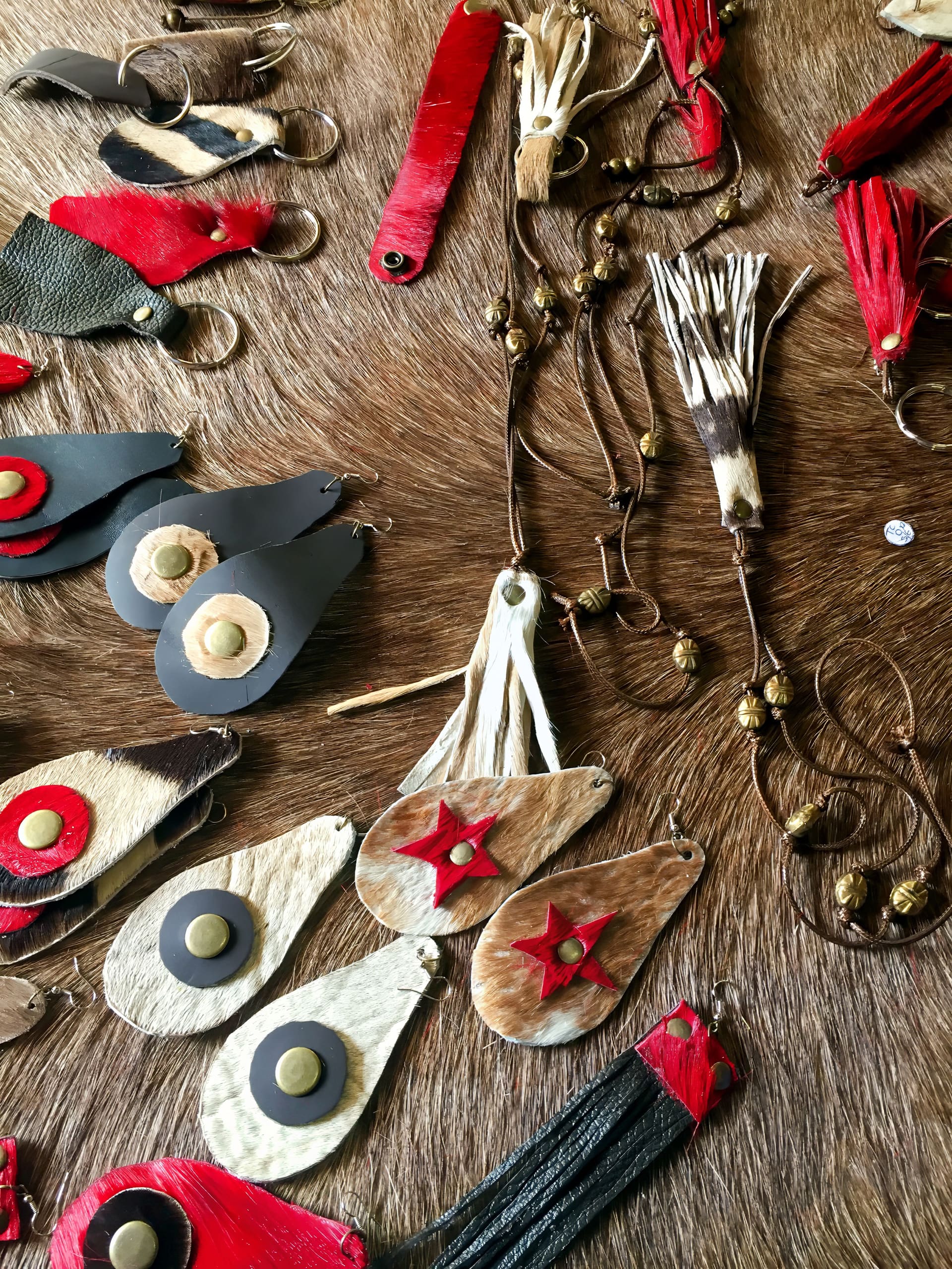 A collection of handmade earrings and necklaces, crafted from leather and fur in red, black, cream, and brown tones, is displayed on a brown furry surface. Designs include teardrops, circles, and stars, some adorned with beads and tassels.