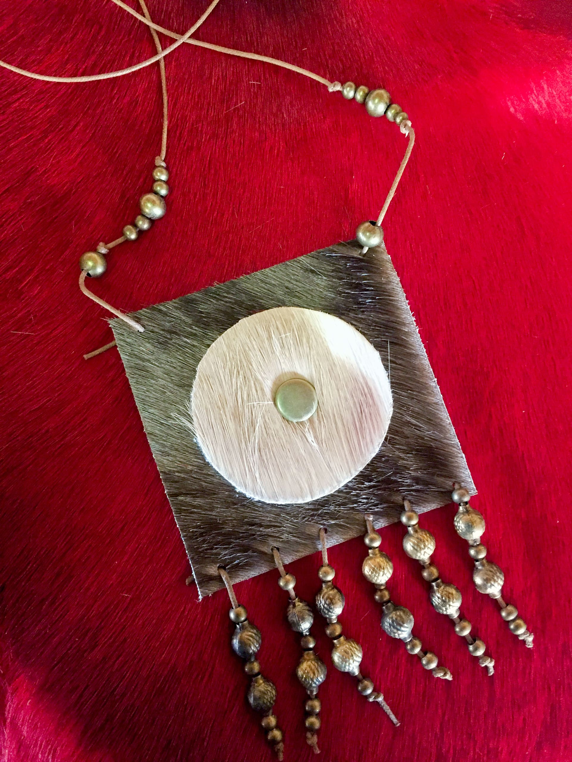 A square pendant necklace with a circular centrepiece and central metal bead, adorned with hanging metal beads in various shapes, is displayed on a deep red textured background. The necklace’s cord features small, round metallic beads.