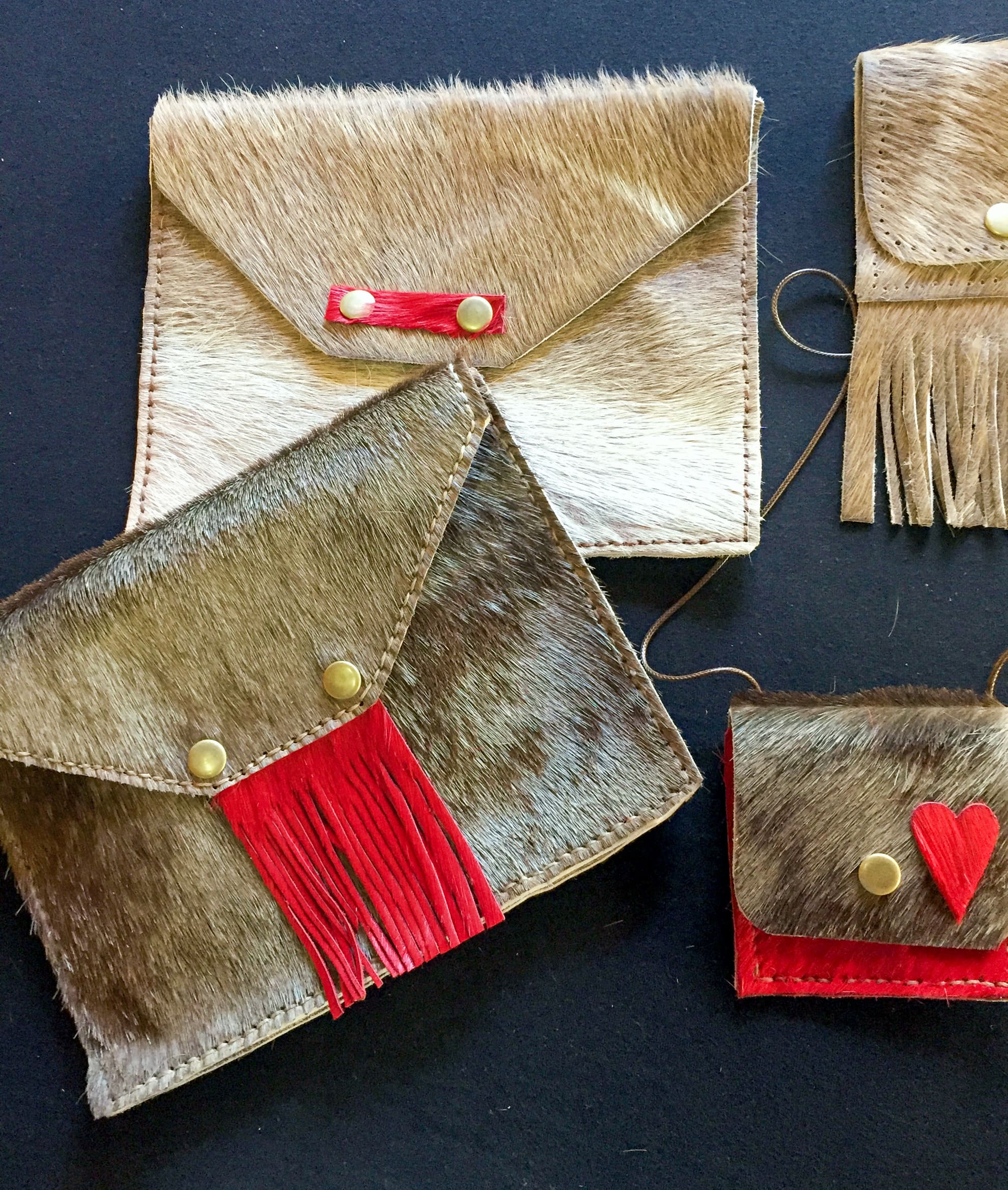 Four hand-sewn envelope-style pouches made of brown and white fur are arranged on a dark surface. Each pouch features gold press studs and red leather details, such as fringe or a heart, adding vibrant accents to the natural textures.