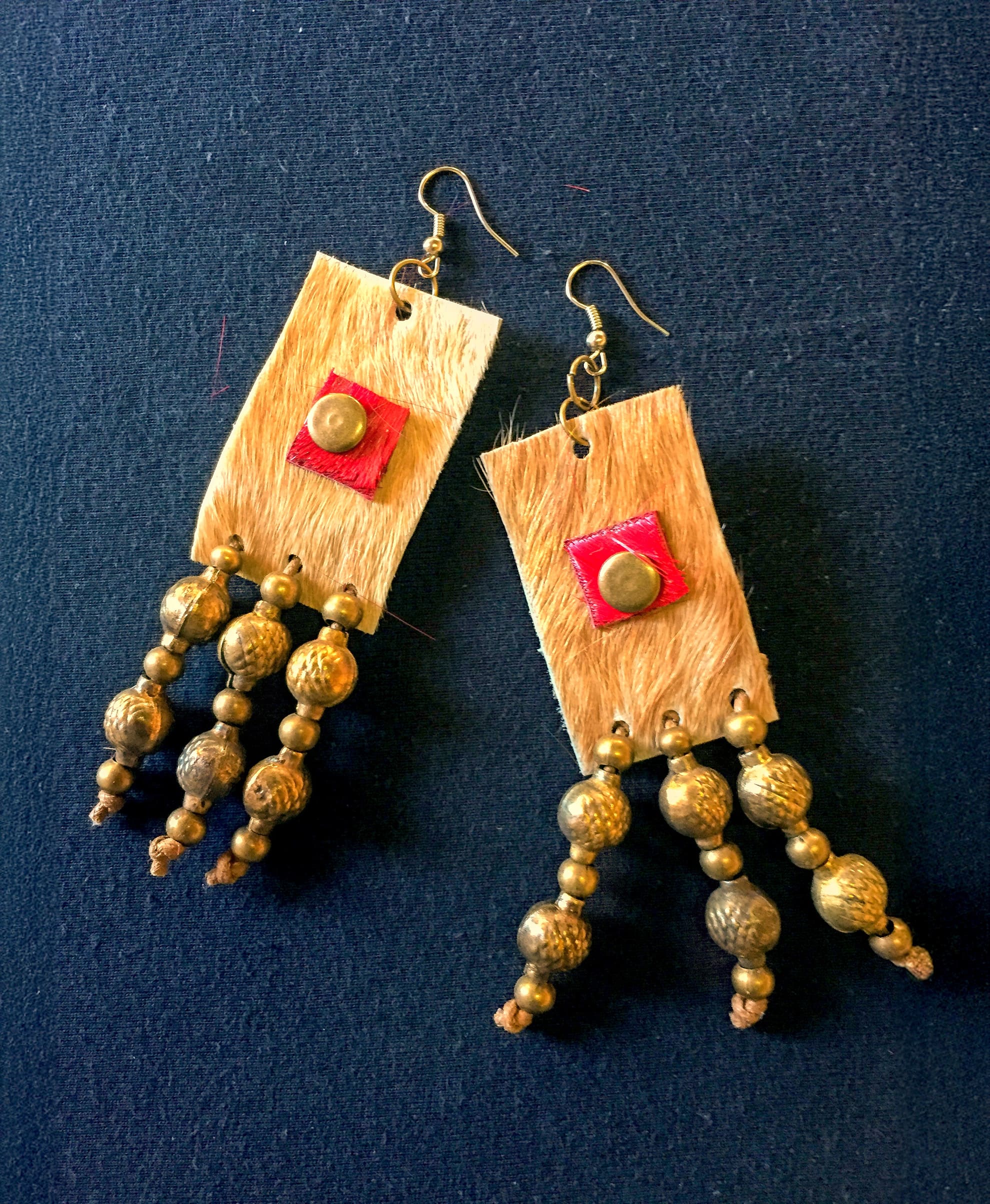 A pair of handmade earrings with rectangular tan fur pieces, each featuring a red square with a round gold stud at the centre. Three strands of textured gold beads dangle from each rectangle. The background is black.