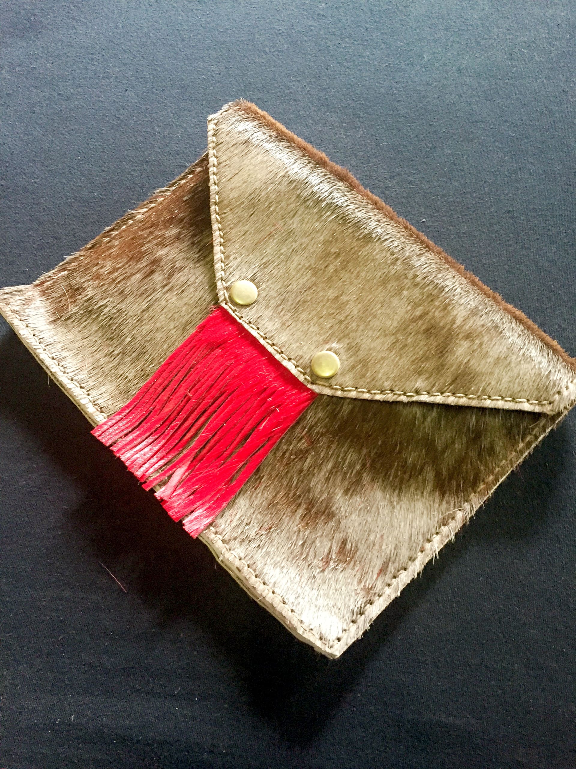 A rectangular brown and beige fur clutch bag with a pointed flap, gold stitching, two gold press studs, and red leather fringe hanging from under the flap. The bag is resting on a dark surface.
