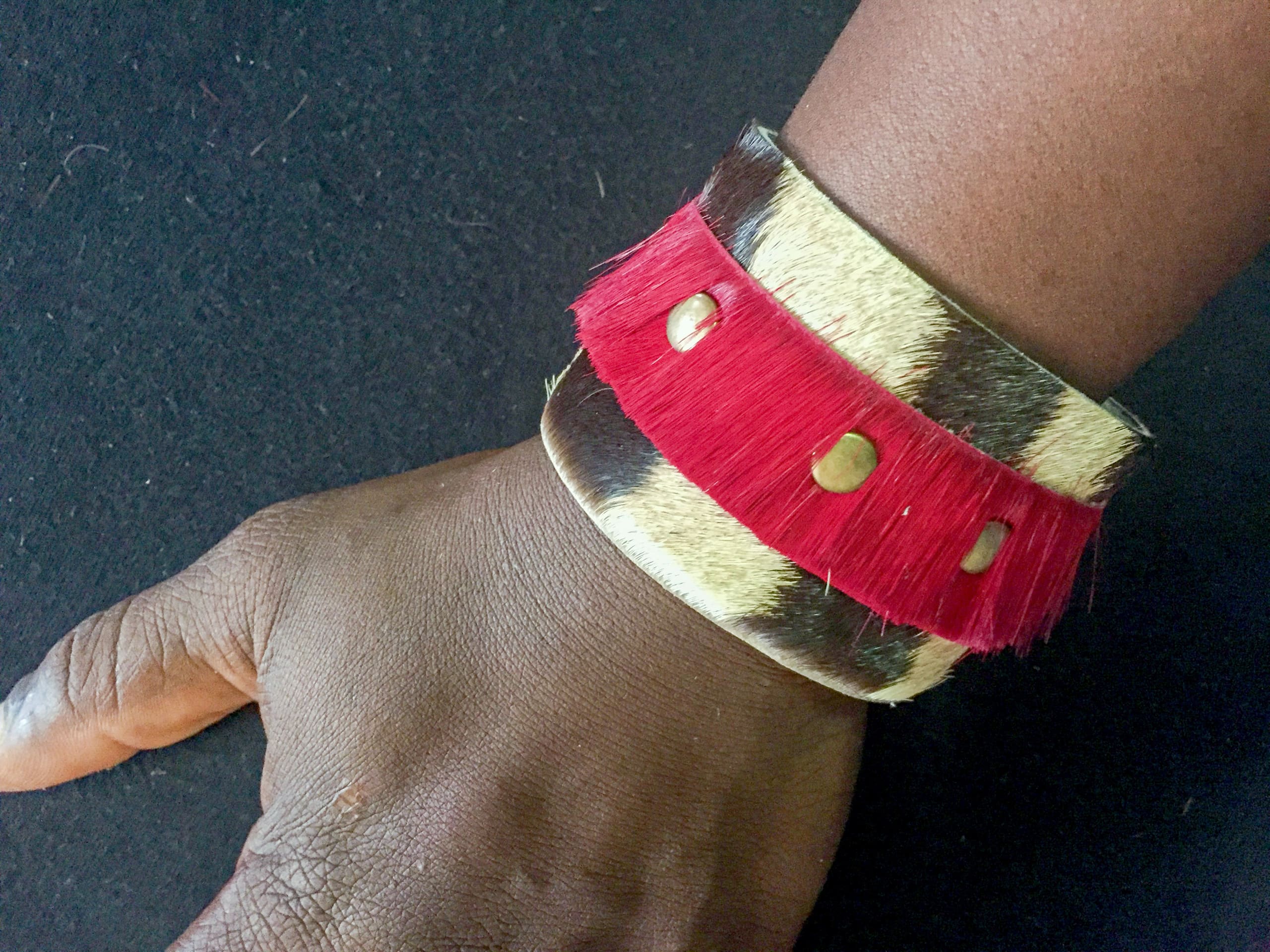 A dark-skinned hand rests on a black surface, wearing a wide, patterned bracelet with an animal print base and a bright red fringe strip. The red band features four brass-like circular studs as accents.