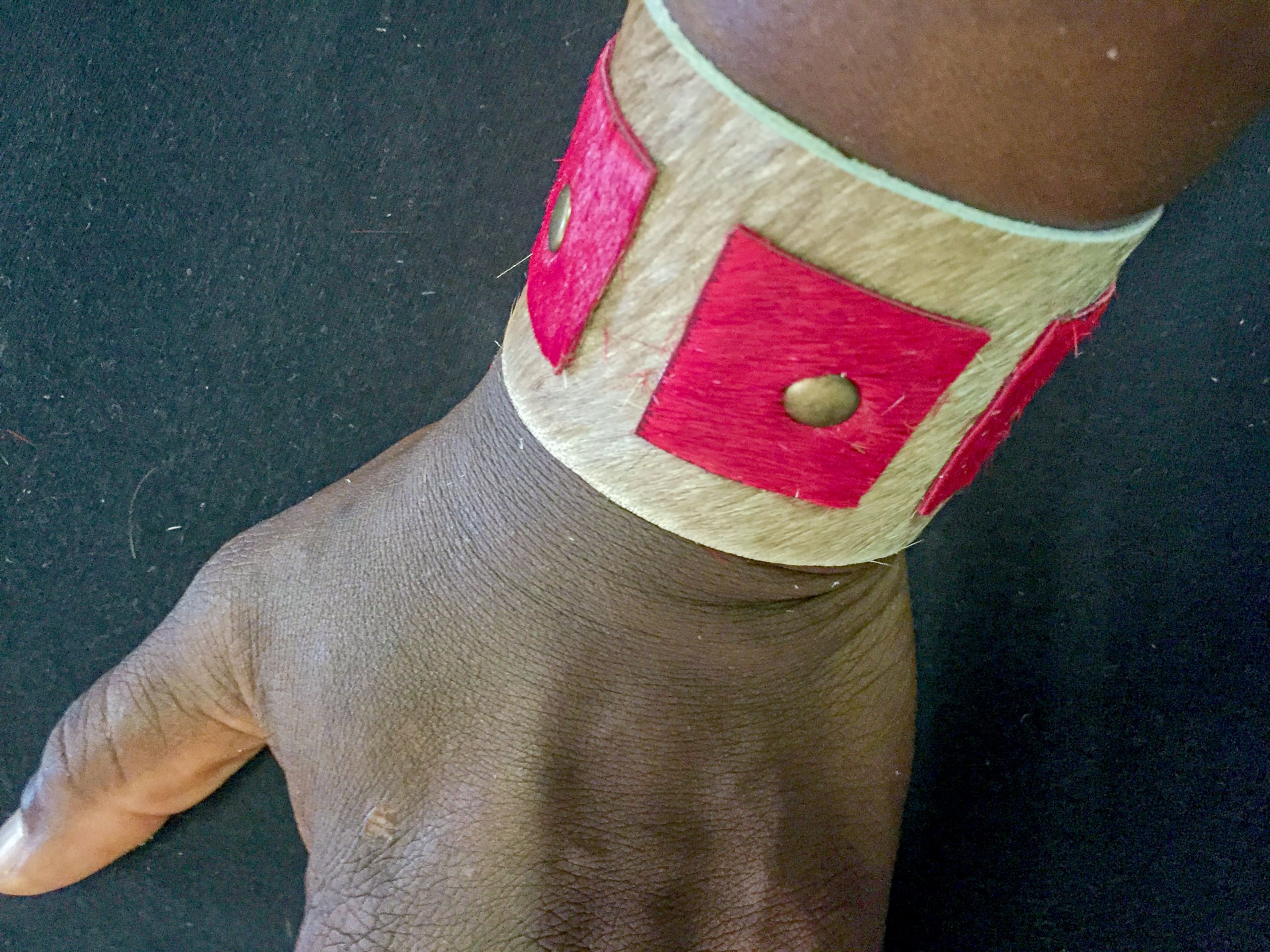 A close-up of a wrist wearing a wide, tan-coloured bracelet with red square decorations and small round metal studs, set against a black background. The bracelet features a textured appearance and contrasts with the wearer’s dark skin tone.
