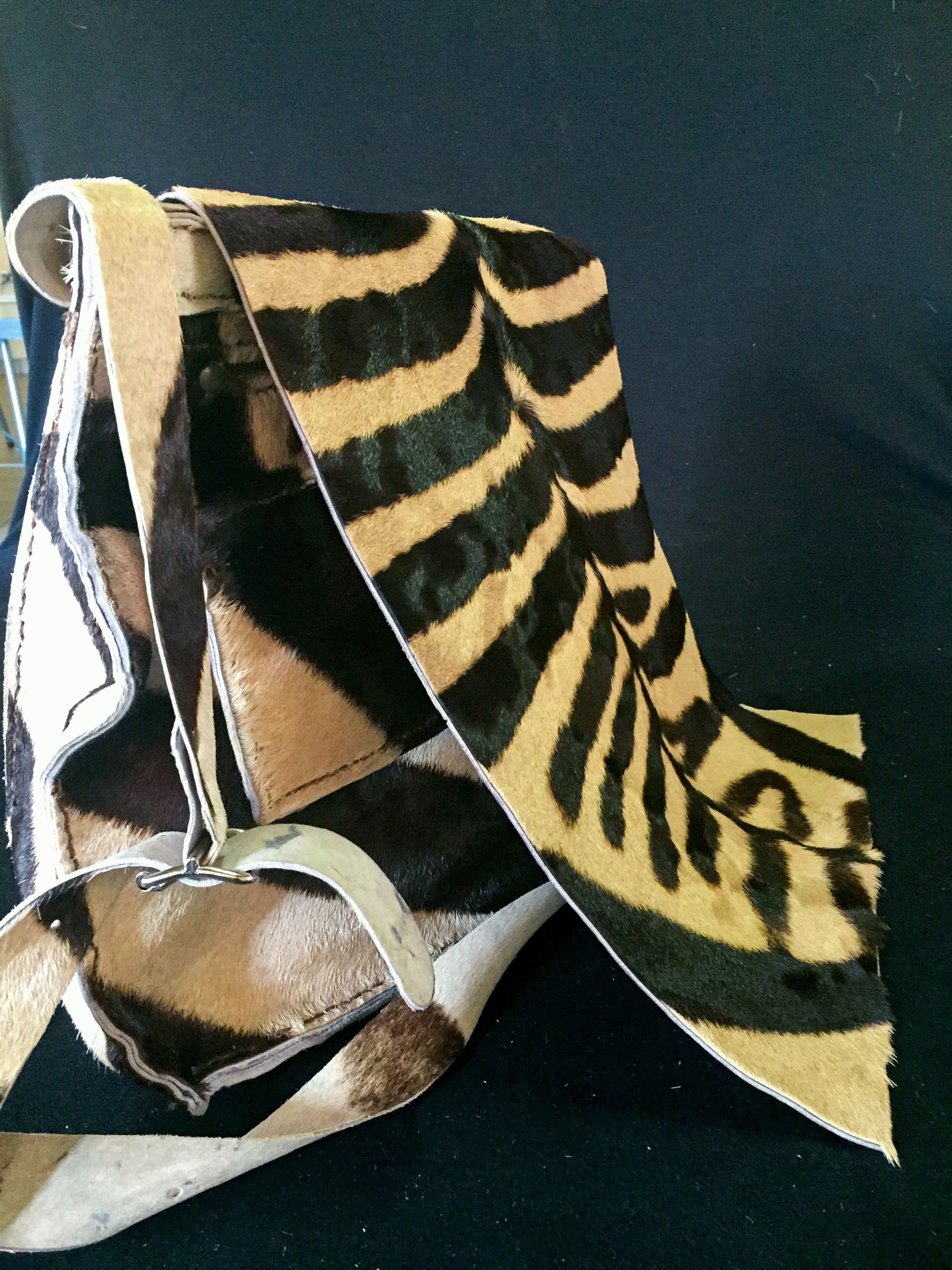 A close-up of a traditional saddle covered with animal hide featuring bold black-and-tan zebra-like stripes, draping over the saddle. The saddle has metal parts and brown-and-white fur accents, and is set against a solid black background.