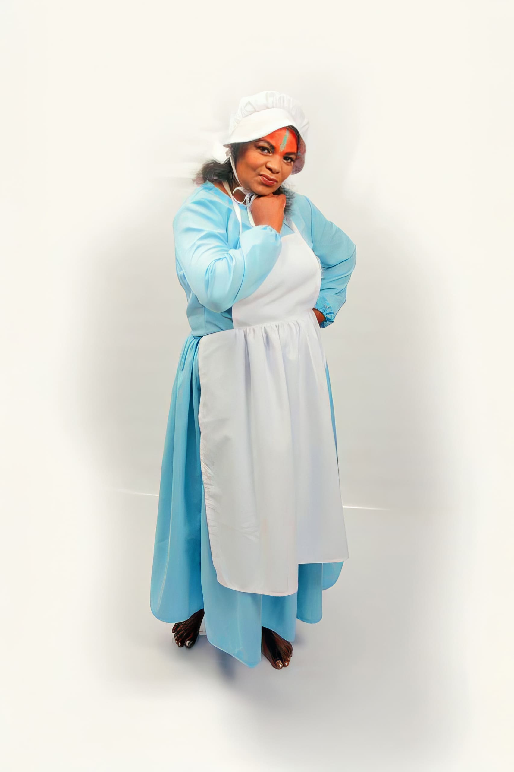 A woman stands barefoot against a white background, wearing a light blue long-sleeved dress, a white apron, and a white bonnet. She poses with one hand under her chin and looks confidently at the camera.