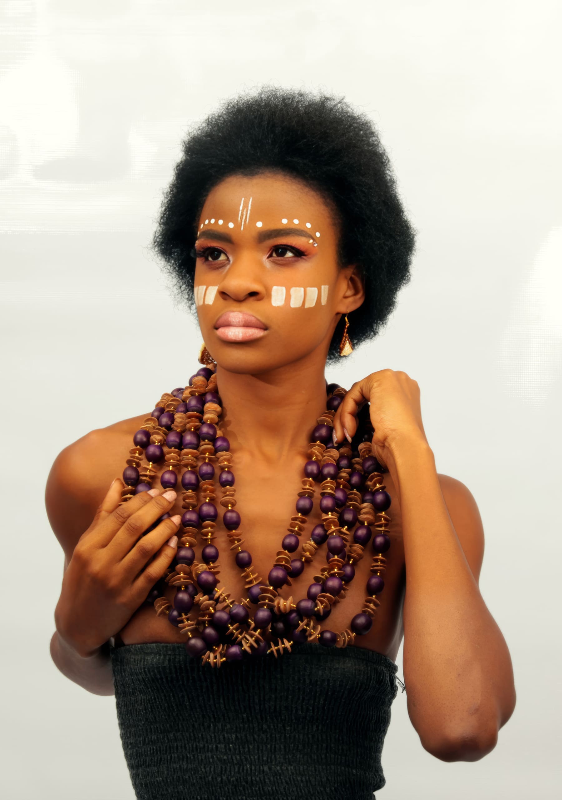 A woman with short natural hair wears tribal face paint—white dots on her forehead and lines on her cheeks. She has layered purple bead necklaces and a black strapless top, posing against a plain light background.