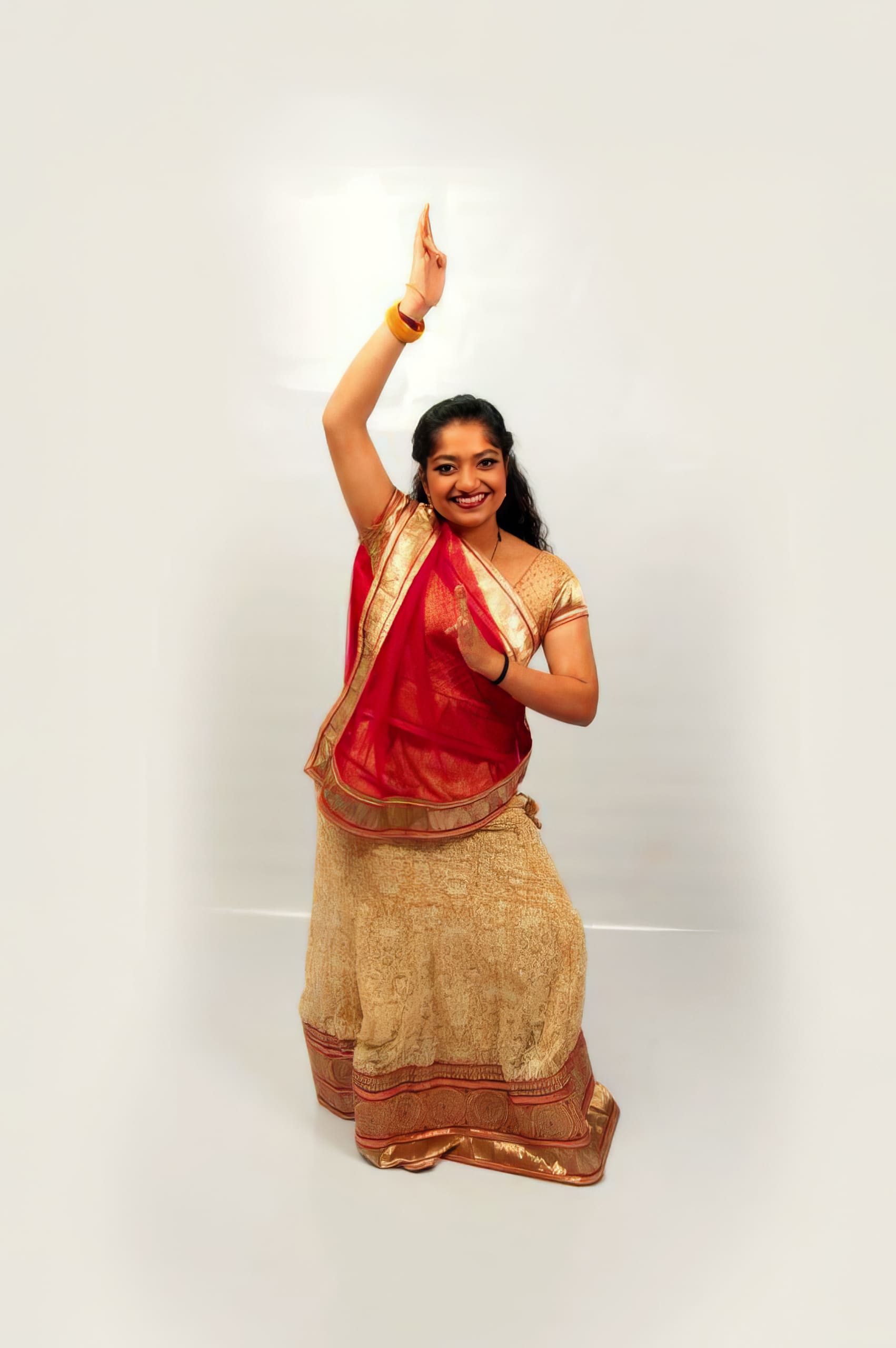 A woman in traditional Indian attire poses in a classical dance stance. She wears a cream and gold lehenga with a red dupatta, gold jewellery, and has long, wavy hair. She smiles whilst raising one arm above her head and bending the other at her waist against a plain background.
