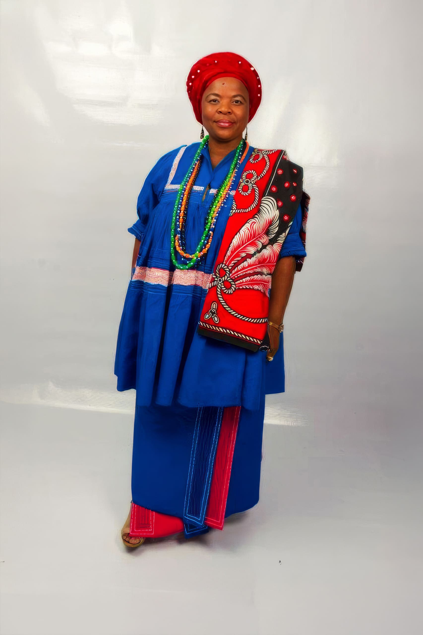 A woman stands against a plain white background, dressed in traditional African attire. She wears a blue dress and headwrap, accessorised with colourful beads and a red patterned shoulder cloth featuring an intricate white and black design. She smiles softly.