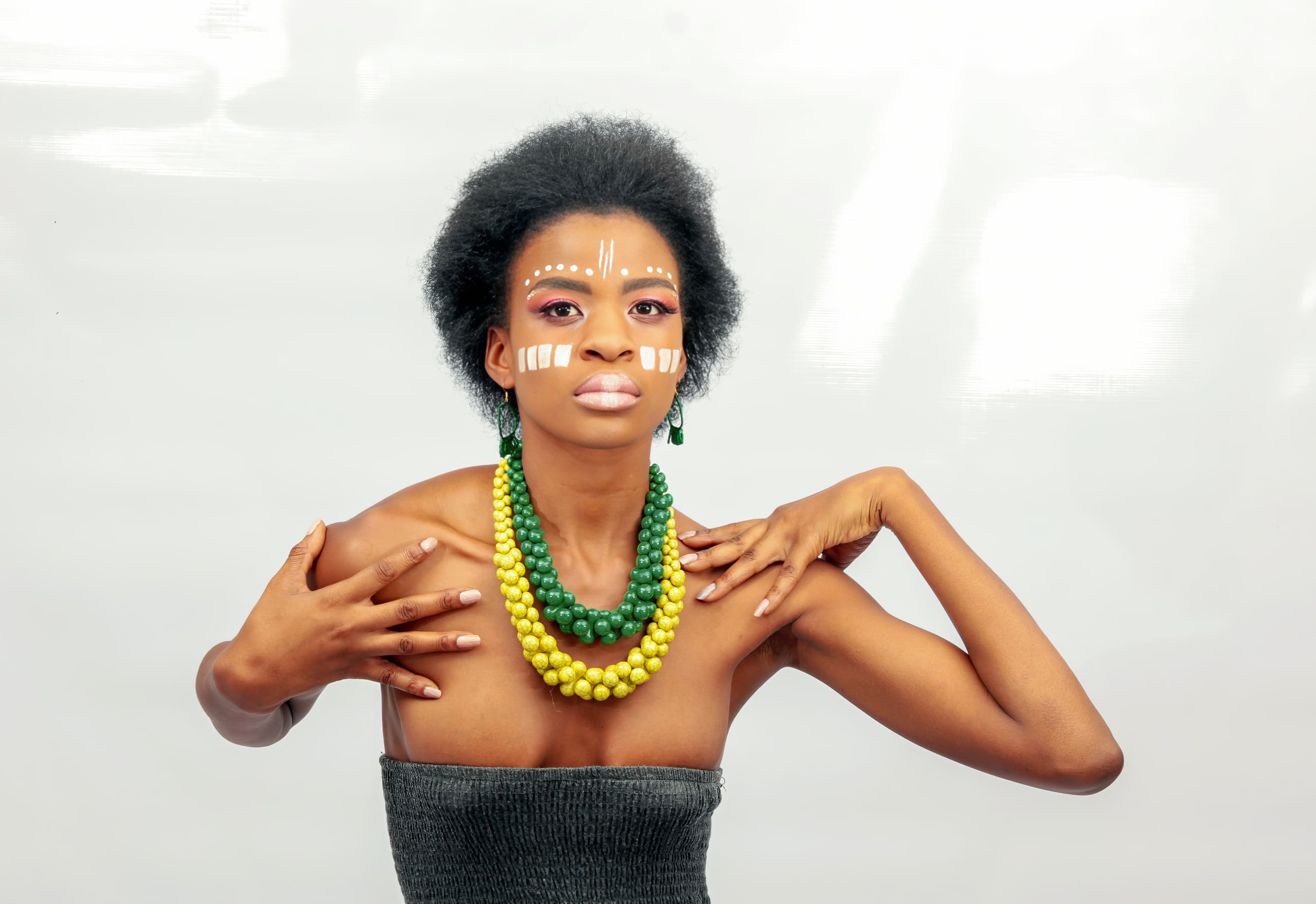 A woman with natural afro hair poses against a white background, wearing yellow and green beaded necklaces and a strapless top. She has bold make-up and white tribal-inspired face paint patterns on her cheeks and forehead. Her hands are raised to her chest and shoulder.