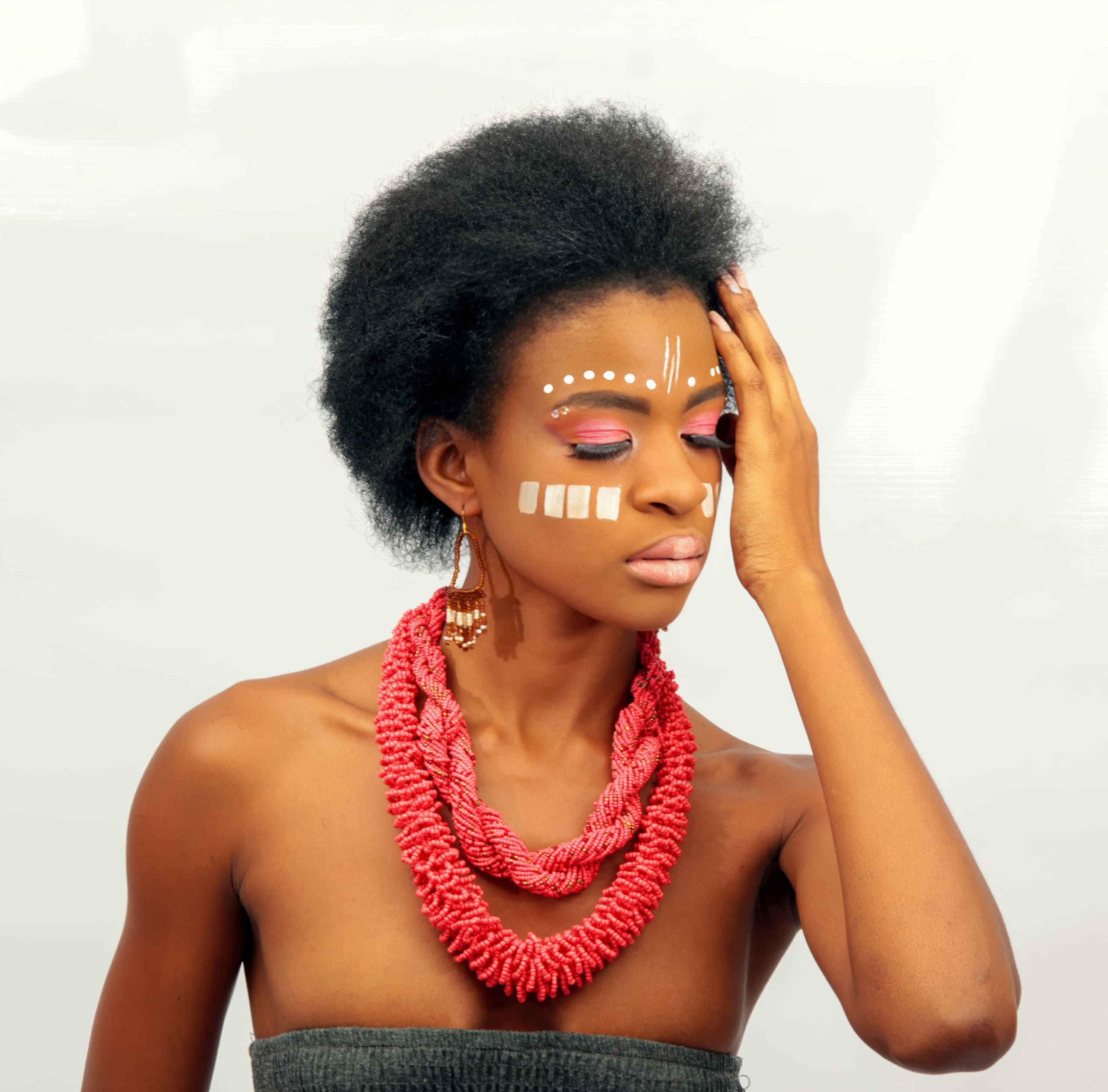 A woman with natural afro hair wears bold face paint in white dots and lines, pink eye make-up, and layered red bead necklaces. She rests one hand on her face with eyes closed, posing against a plain light background.
