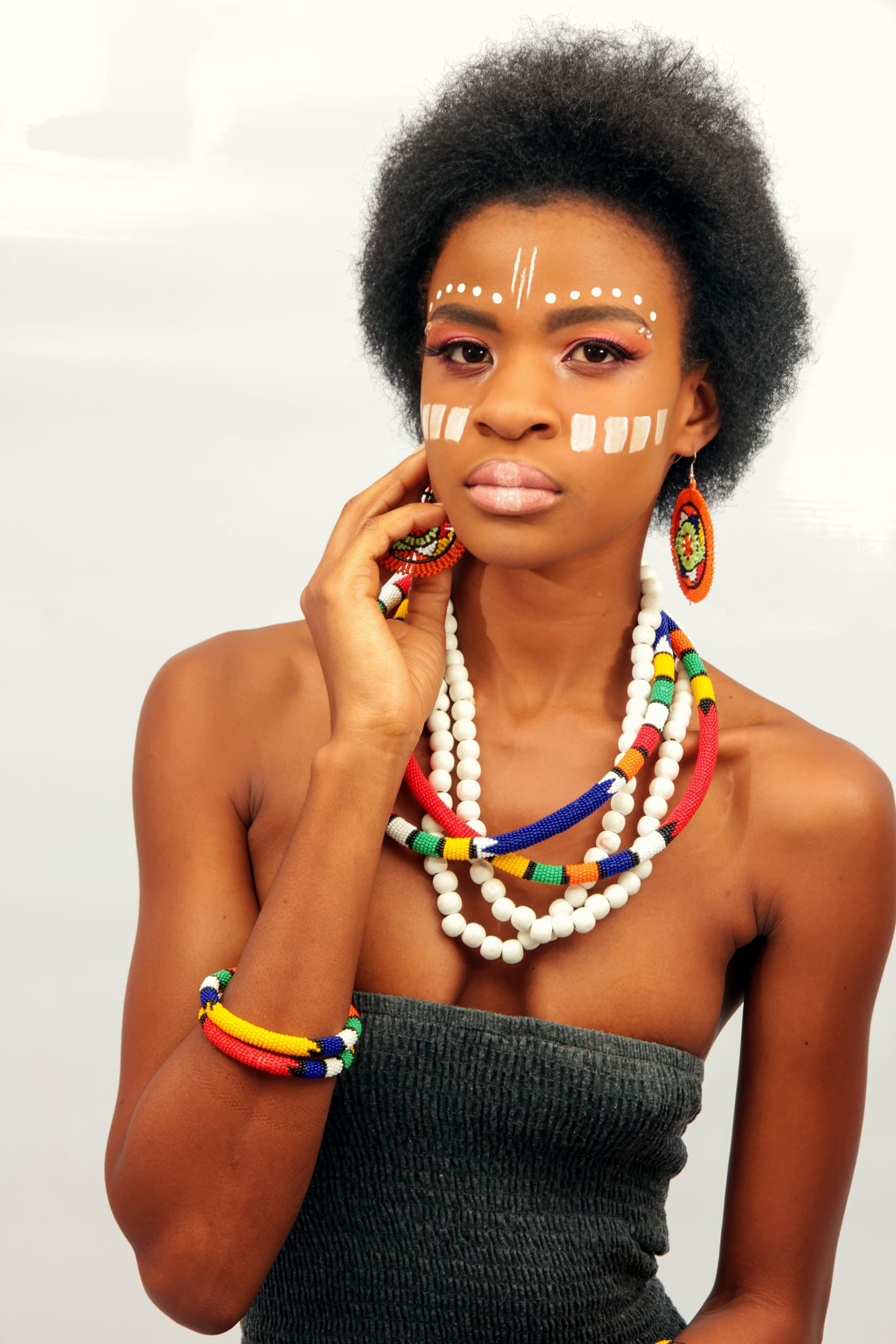 A woman with short, natural hair wears face paint with white dots and lines, colourful beaded jewellery, and a strapless dark top. She gazes at the camera with one hand touching her face. The background is plain and light, creating a striking portrait.