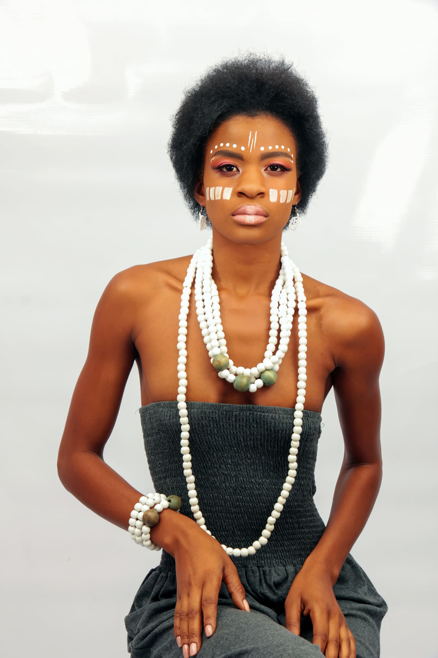 A woman with short curly hair wears white and brown face paint in geometric patterns. She is dressed in a strapless dark grey top, adorned with layered white bead necklaces and a matching bracelet. She sits upright against a plain light background.
