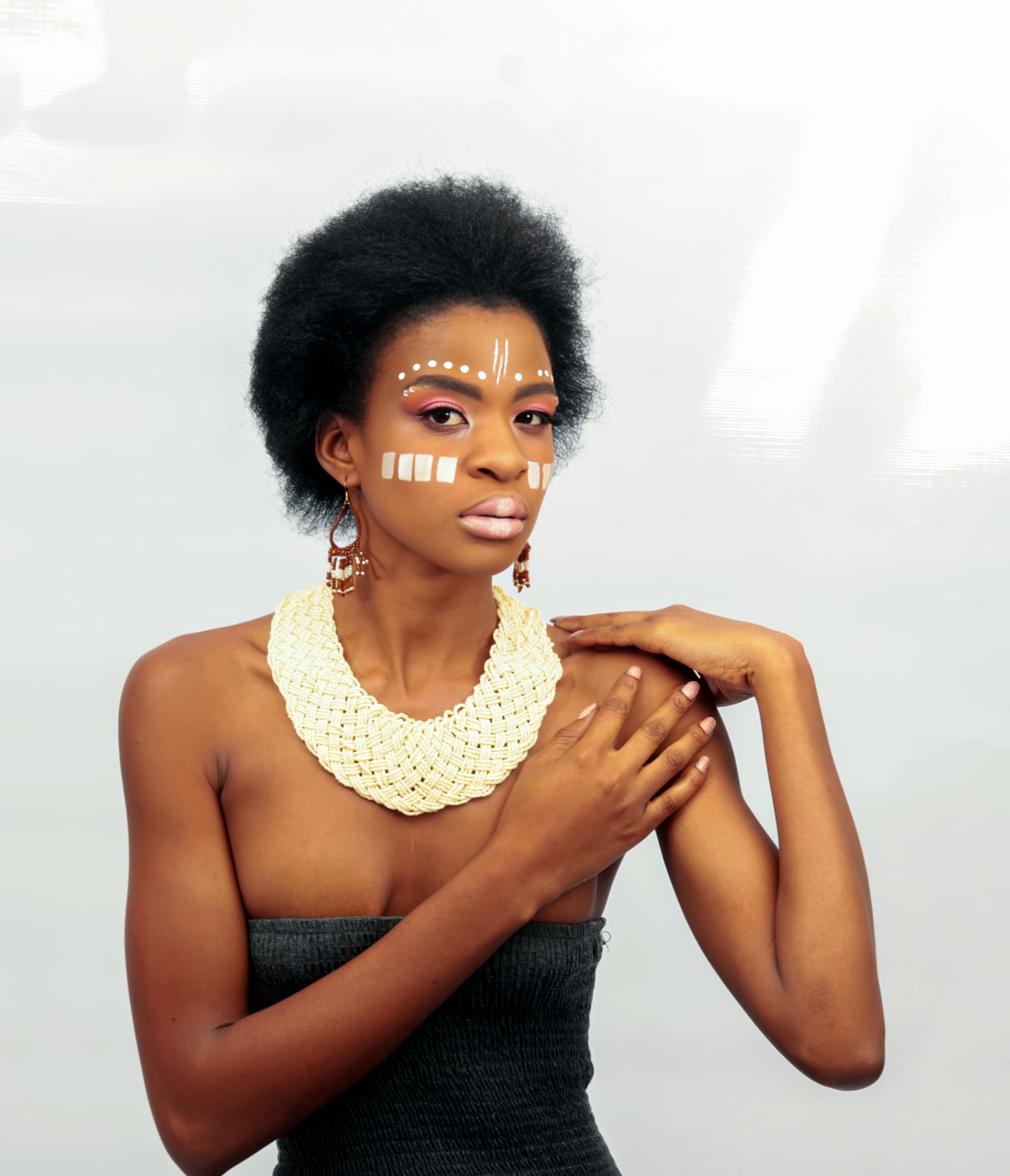 A woman with natural afro hair poses against a light background. She wears a strapless black top, a chunky cream braided necklace, and brown earrings. Her face is decorated with white tribal paint and her make-up includes pink eyeshadow and lipstick.