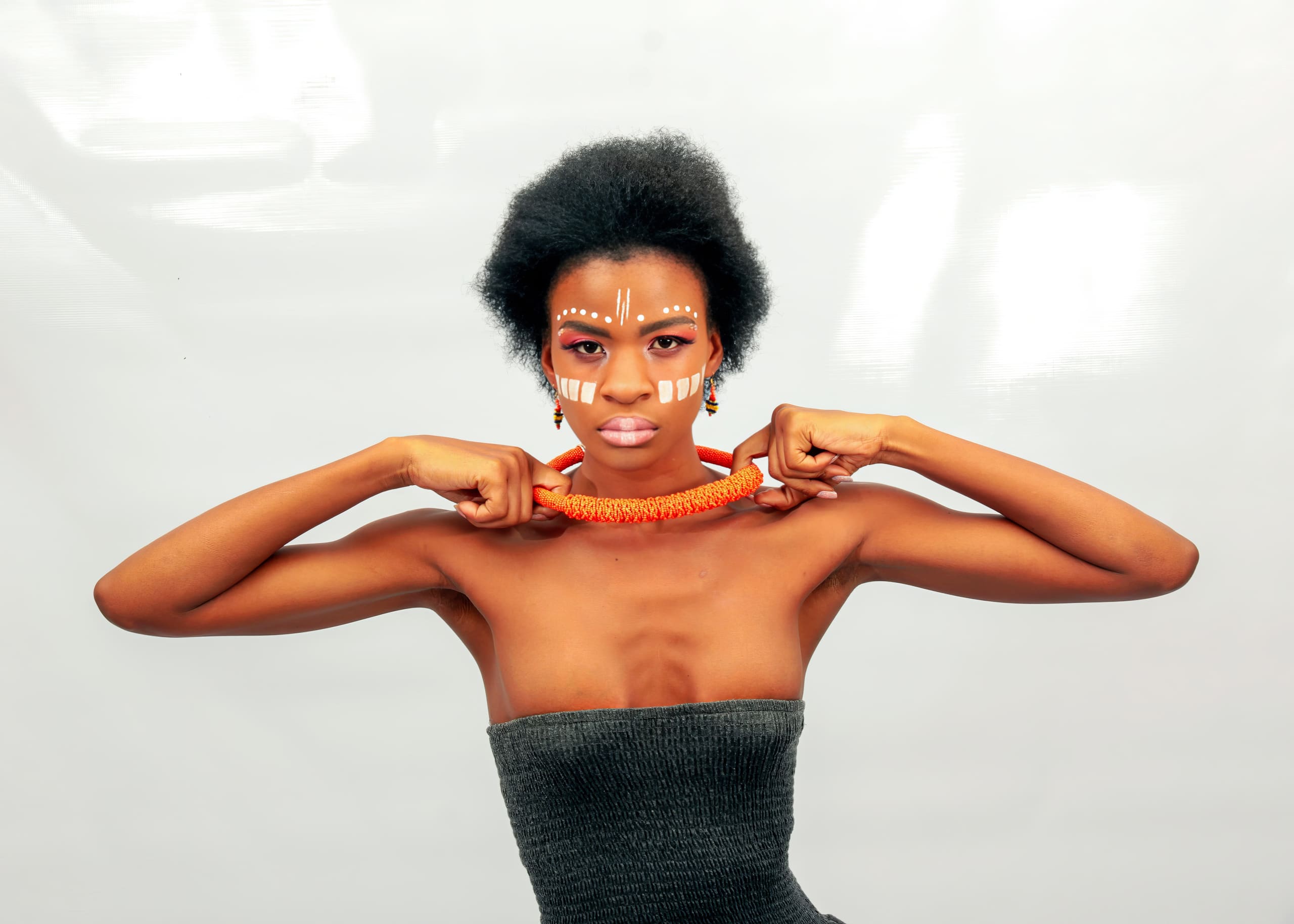 A woman with short natural hair, wearing a strapless grey top and orange beaded necklace, poses with both hands holding the necklace. She has white tribal face paint and orange earrings, standing against a plain light background.