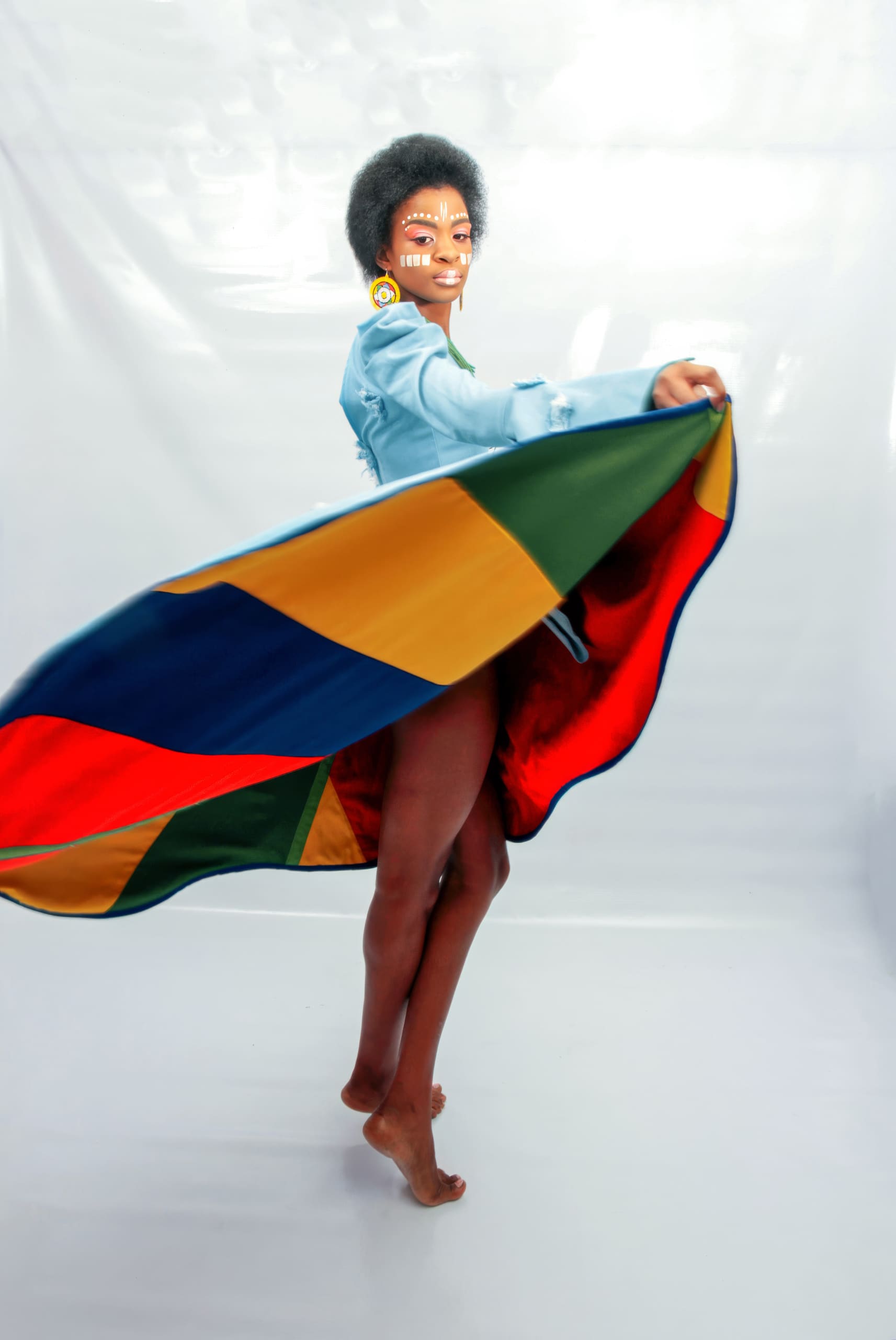 A barefoot person with face paint, earrings, and an afro hairstyle twirls, causing a colourful, geometric-patterned skirt to fan out. The background is plain white, highlighting the movement and vivid colours of the skirt.