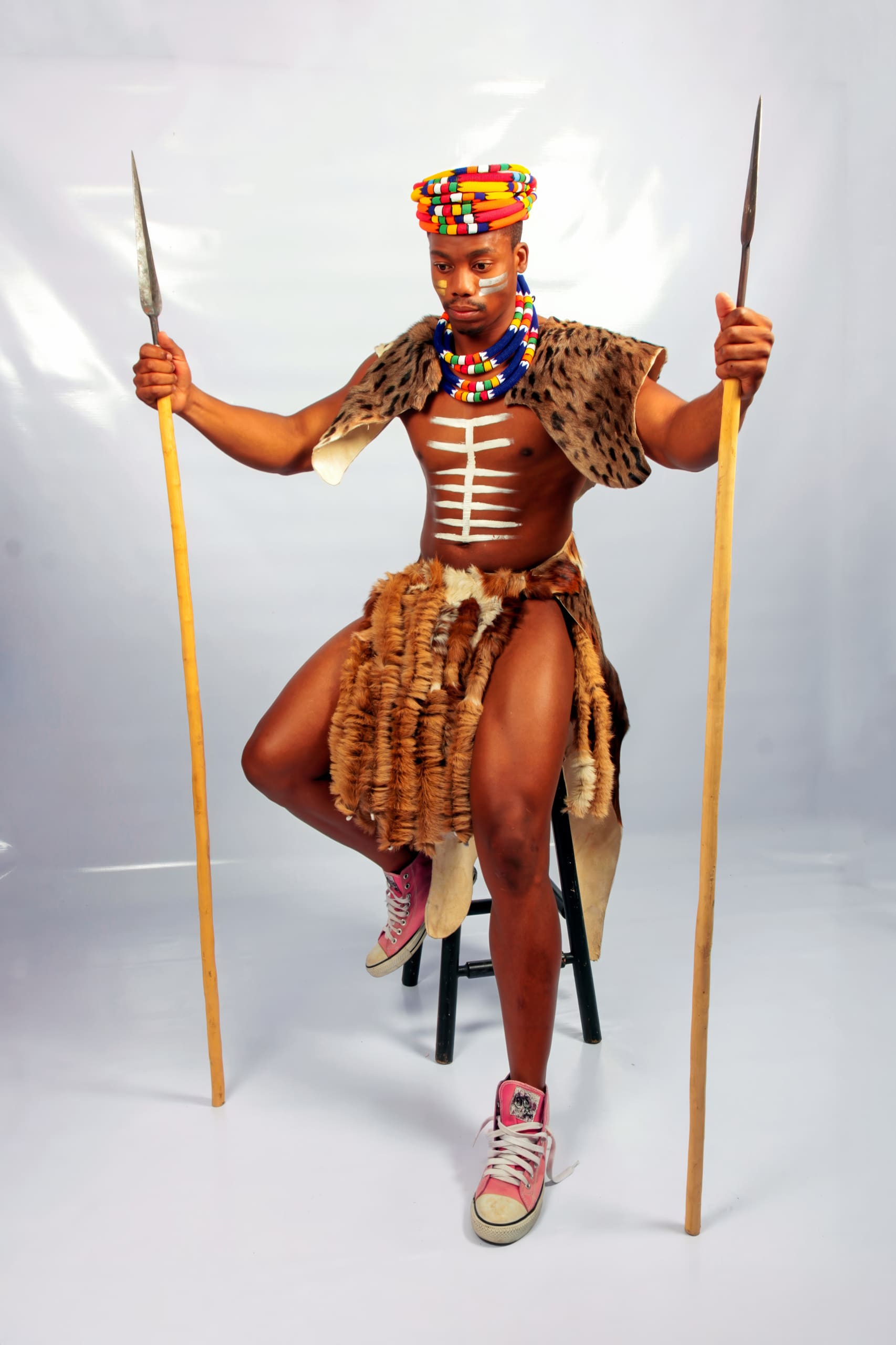 A man wearing traditional African attire with animal fur, colourful beaded jewellery, and a matching headpiece sits on a stool. He holds a spear in each hand and wears pink Converse trainers. The background is plain white.