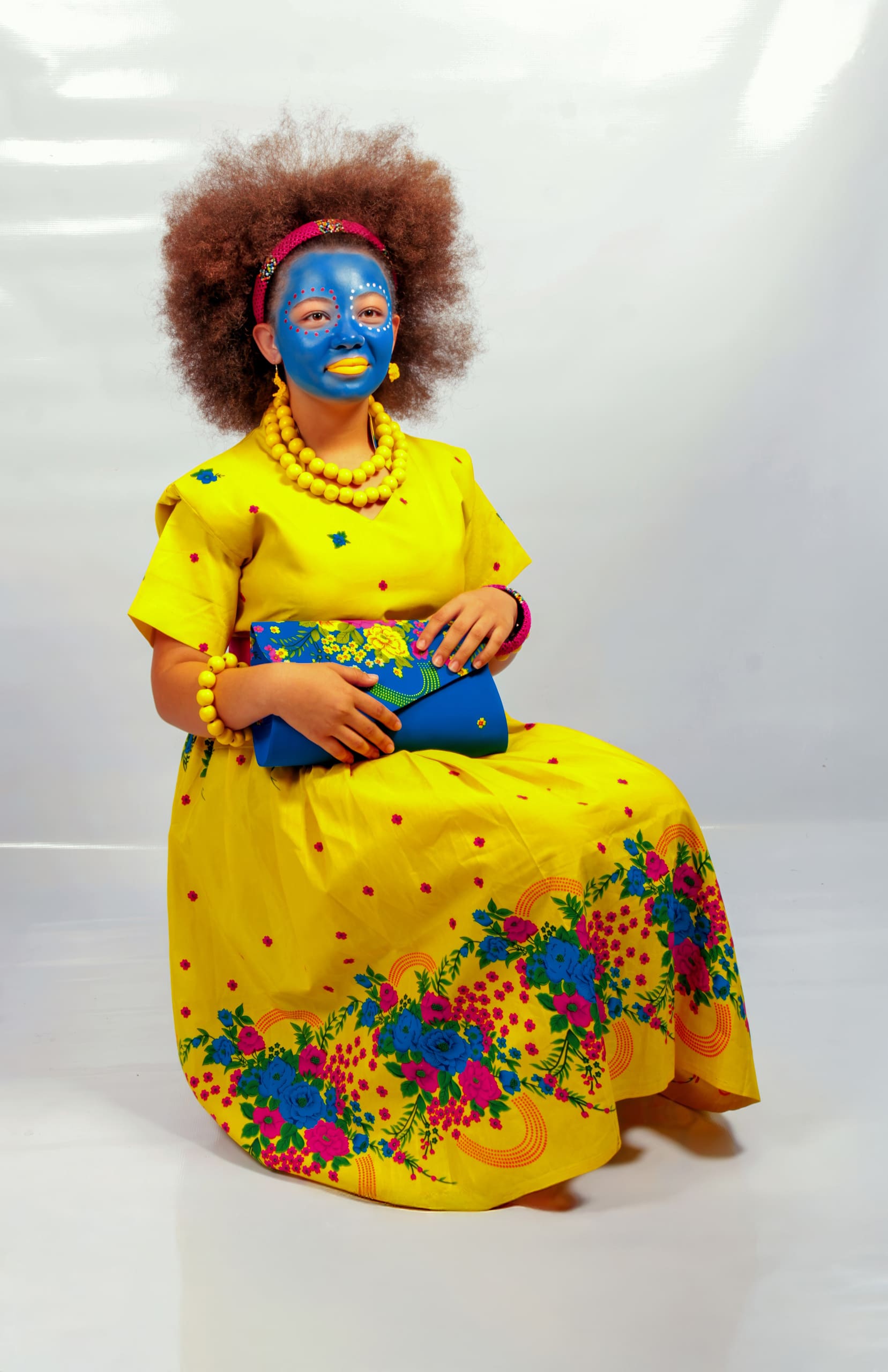 A person with a large afro and blue-painted face sits on a stool, wearing a bright yellow dress with colourful floral patterns, matching yellow jewellery, and holding a blue clutch. The background is plain white, highlighting their vibrant attire.