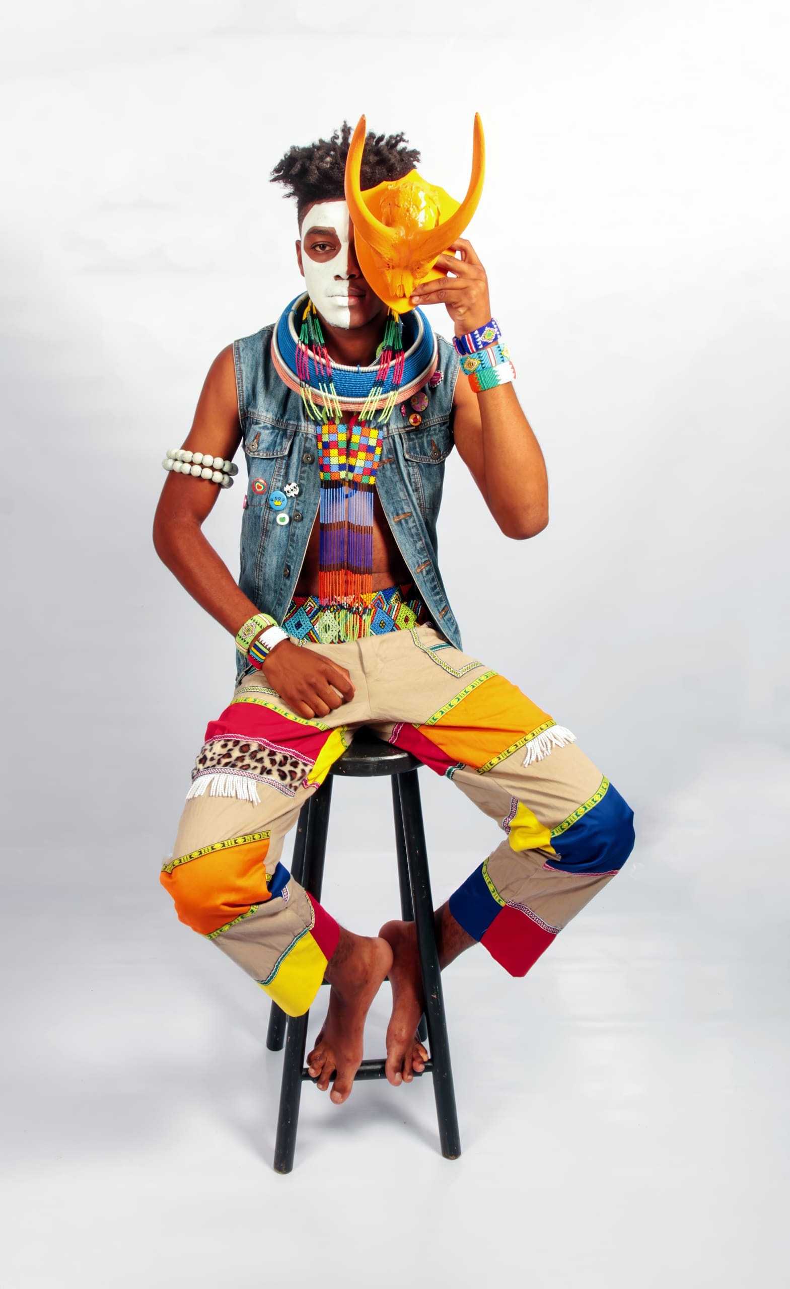 A barefoot person sits on a stool wearing colourful patchwork trousers, a denim waistcoat, layered beaded necklaces, and bracelets. Their face is painted white and black, and they hold a bright yellow horned mask over part of their face against a light background.