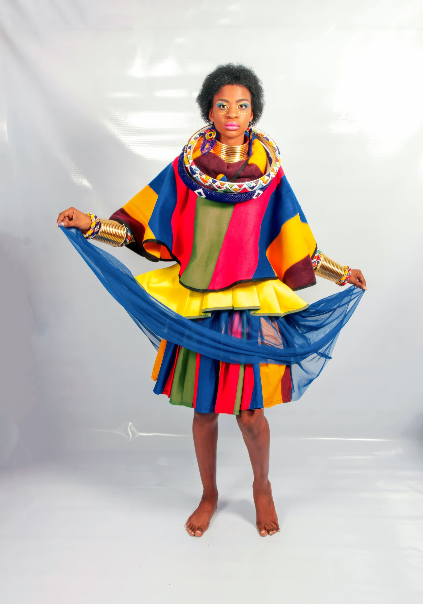 A barefoot woman stands on a white background wearing a colourful, patterned outfit with bold stripes of red, yellow, green, and blue, a layered skirt, gold bangles, and a beaded necklace. She holds out a sheer blue scarf, displaying traditional African attire.