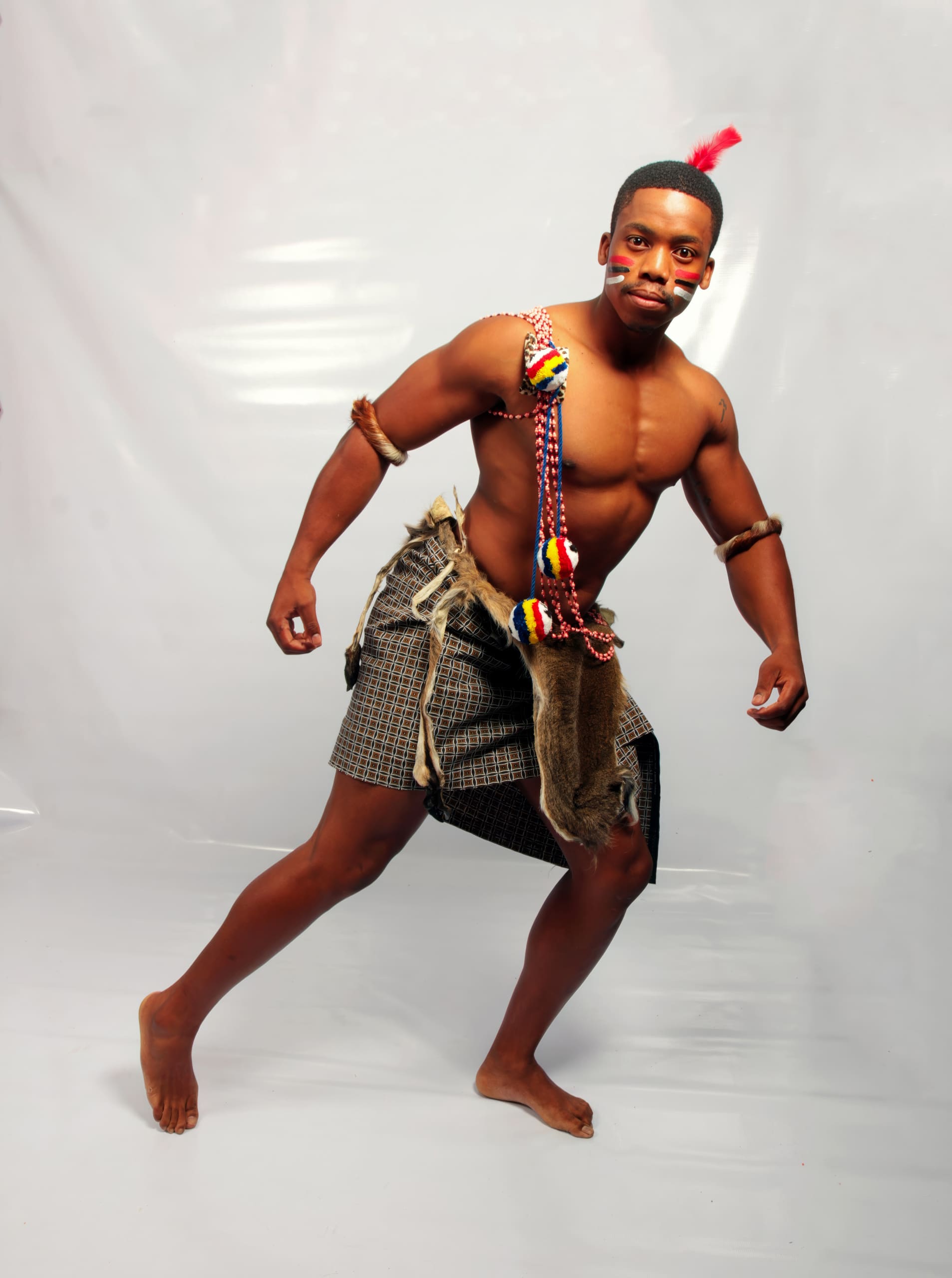 A barefoot man poses with a strong stance, wearing traditional patterned shorts, fur, and colourful bead accessories across his chest and arms. He has tribal face paint and a small red feather in his hair, set against a plain light background.