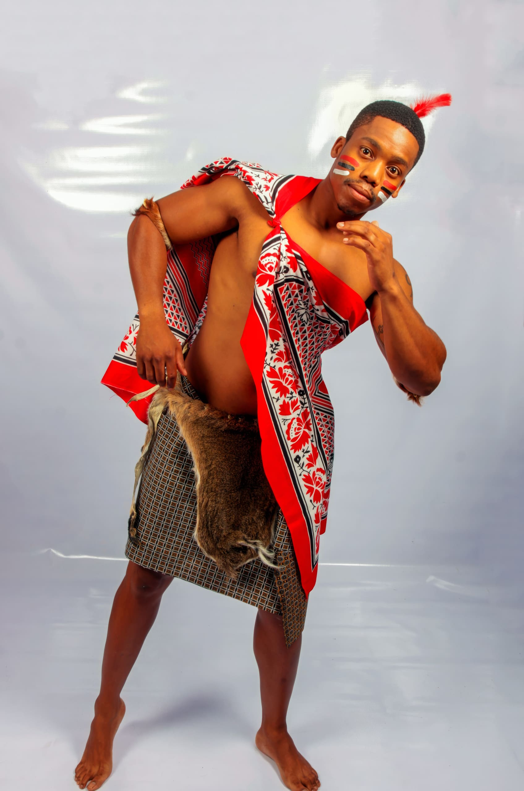A barefoot man poses wearing traditional Swazi attire: a red, black, and white patterned cloth draped over one shoulder and a wrap skirt with fur. He has a feather headpiece and white face paint, standing against a plain, light-coloured background.