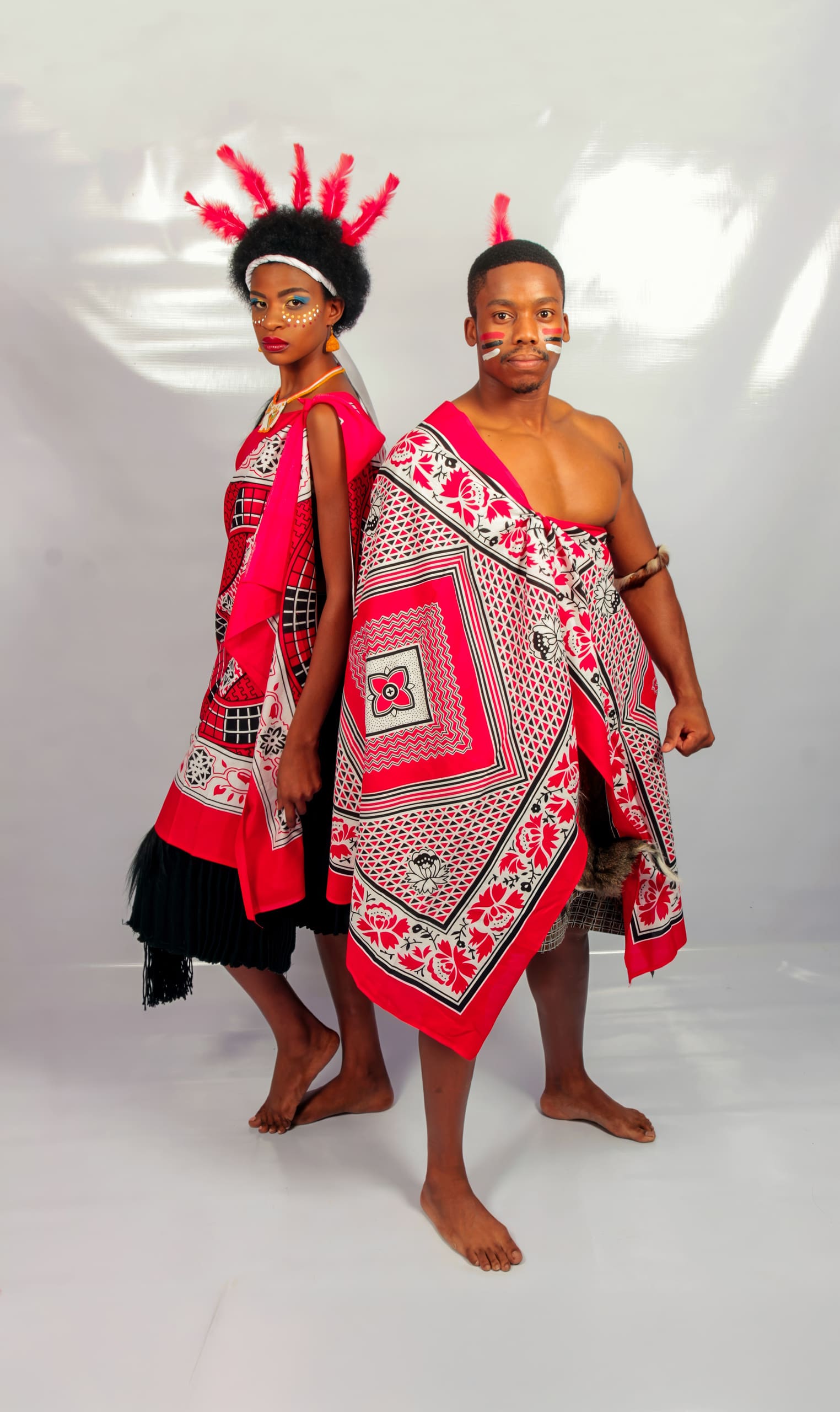 A woman and a man stand back to back wearing vibrant red, black, and white traditional attire with geometric patterns. The woman has a decorated headdress with red feathers and painted face; the man is bare-chested with a patterned wrap and painted cheeks.