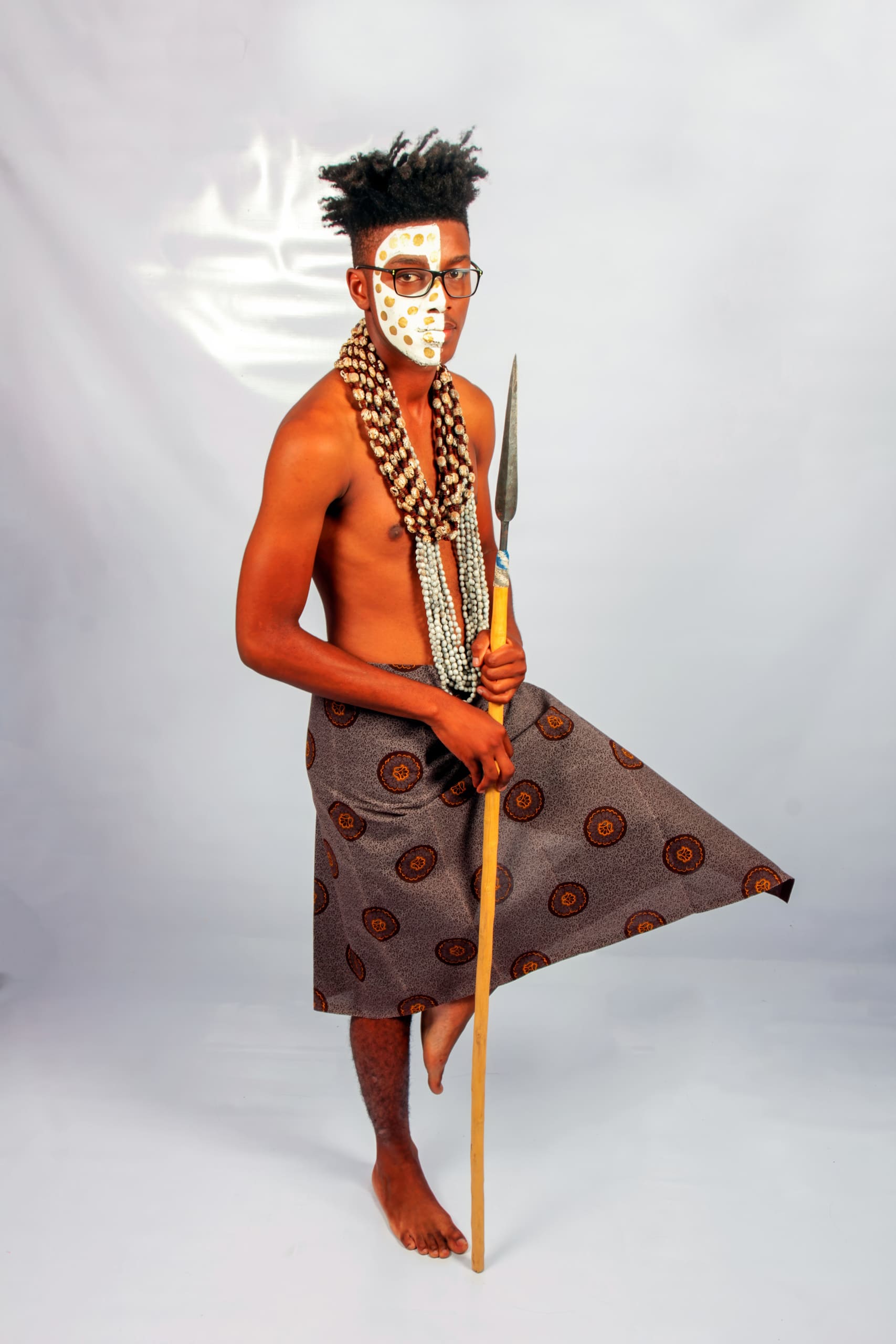 A barefoot man stands on one leg against a plain backdrop, wearing a patterned wrap skirt, layered beaded necklaces, and traditional white face paint. He holds a spear in one hand and gazes forward, embodying traditional African attire and cultural heritage.