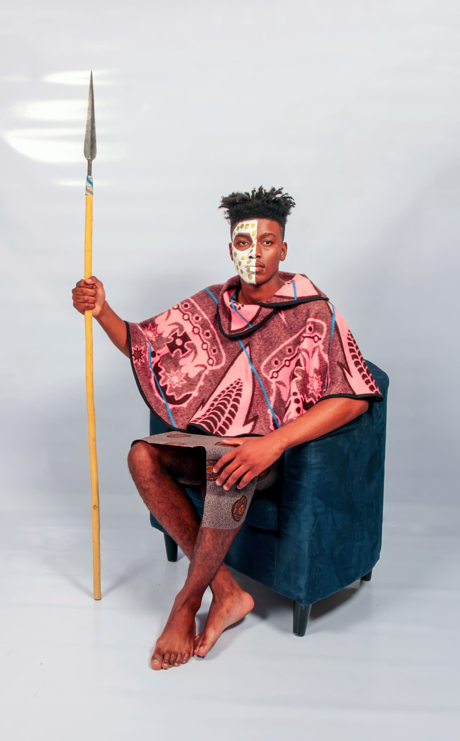 A barefoot man sits on a blue chair, wearing a patterned pink and brown poncho and face paint. He holds a long spear upright in his right hand, and his left knee is bandaged. The background is plain and light-coloured.