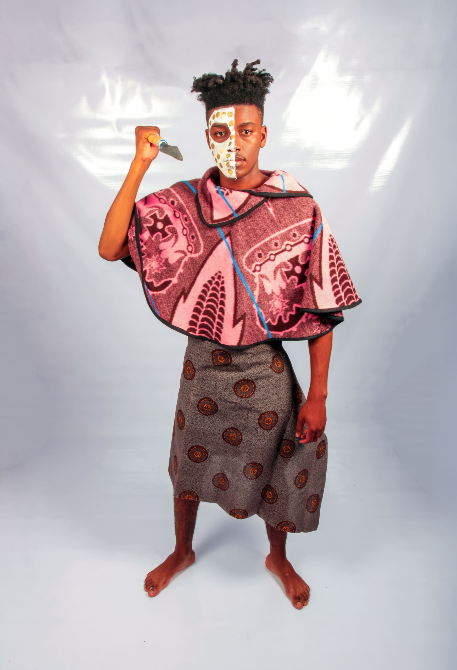A barefoot person stands on a light floor, wearing patterned traditional clothing: a pink-and-purple poncho and a grey skirt with circular designs. They hold a knife upright, and half their face is painted white with distinct markings. The background is plain and light-coloured.