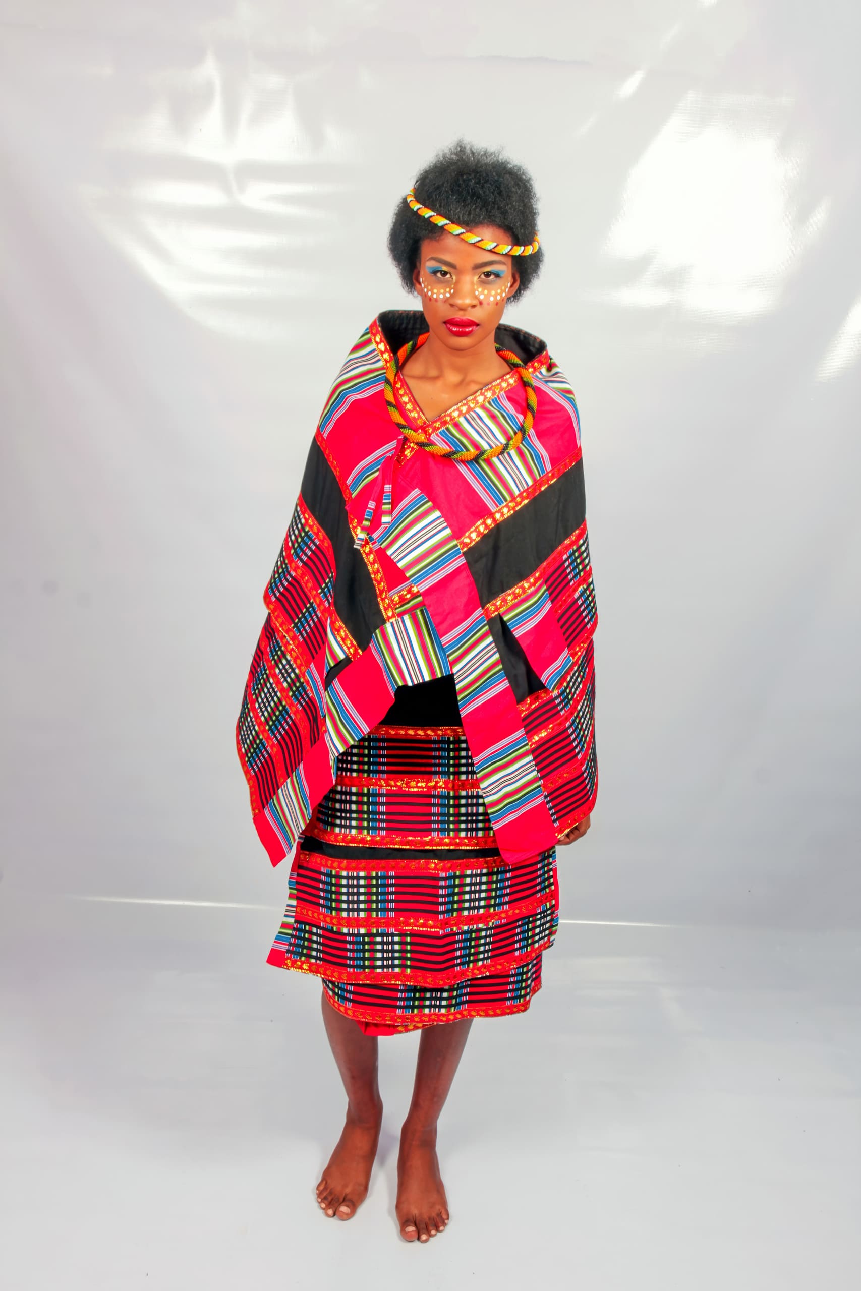 A woman with natural hair stands barefoot against a plain backdrop, wearing traditional African attire with bold red, black, and multicoloured geometric patterns. She has face paint, a beaded headband, and a shawl draped over her shoulders and skirt.