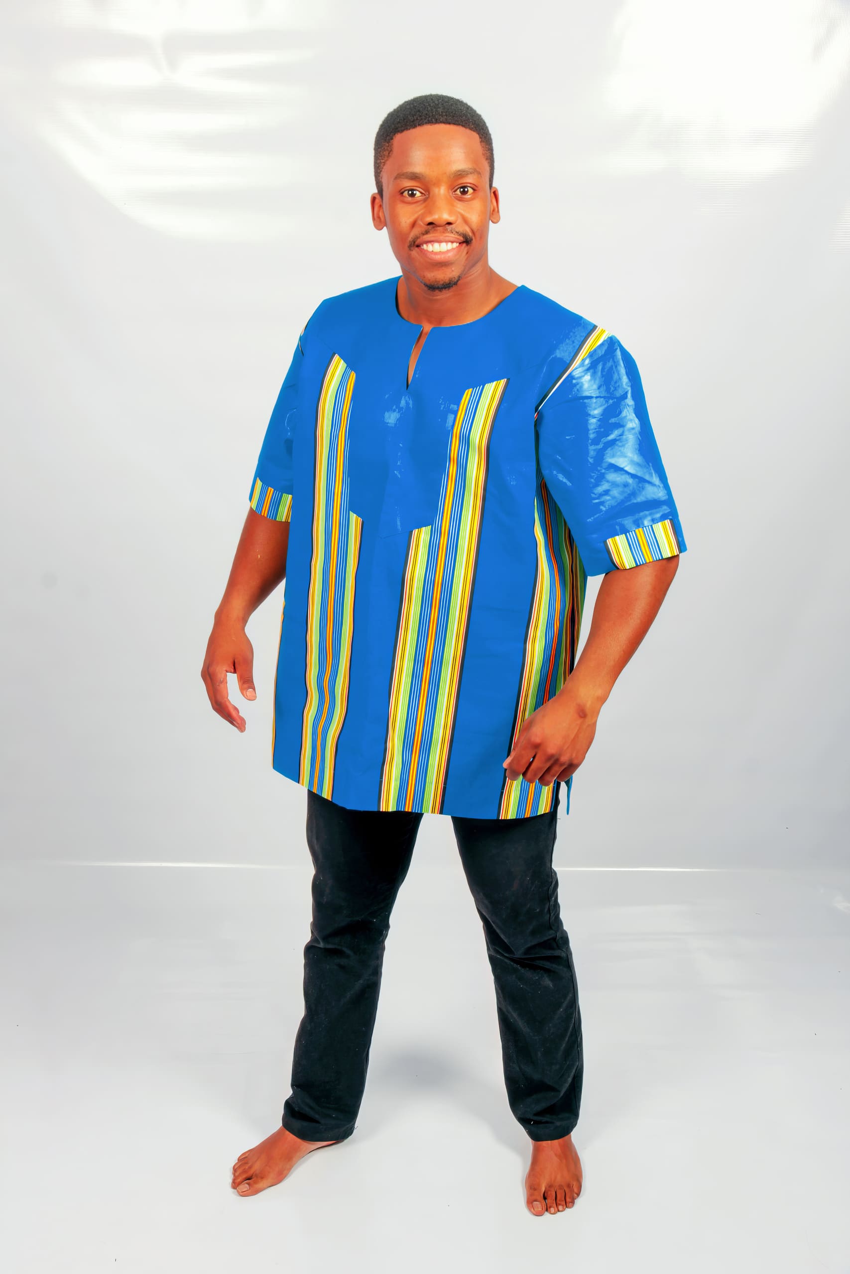 A smiling man stands barefoot on a white floor against a light background. He wears a blue shirt with yellow, green, and white vertical stripes and black trousers. His arms are relaxed by his sides, and he faces the camera.