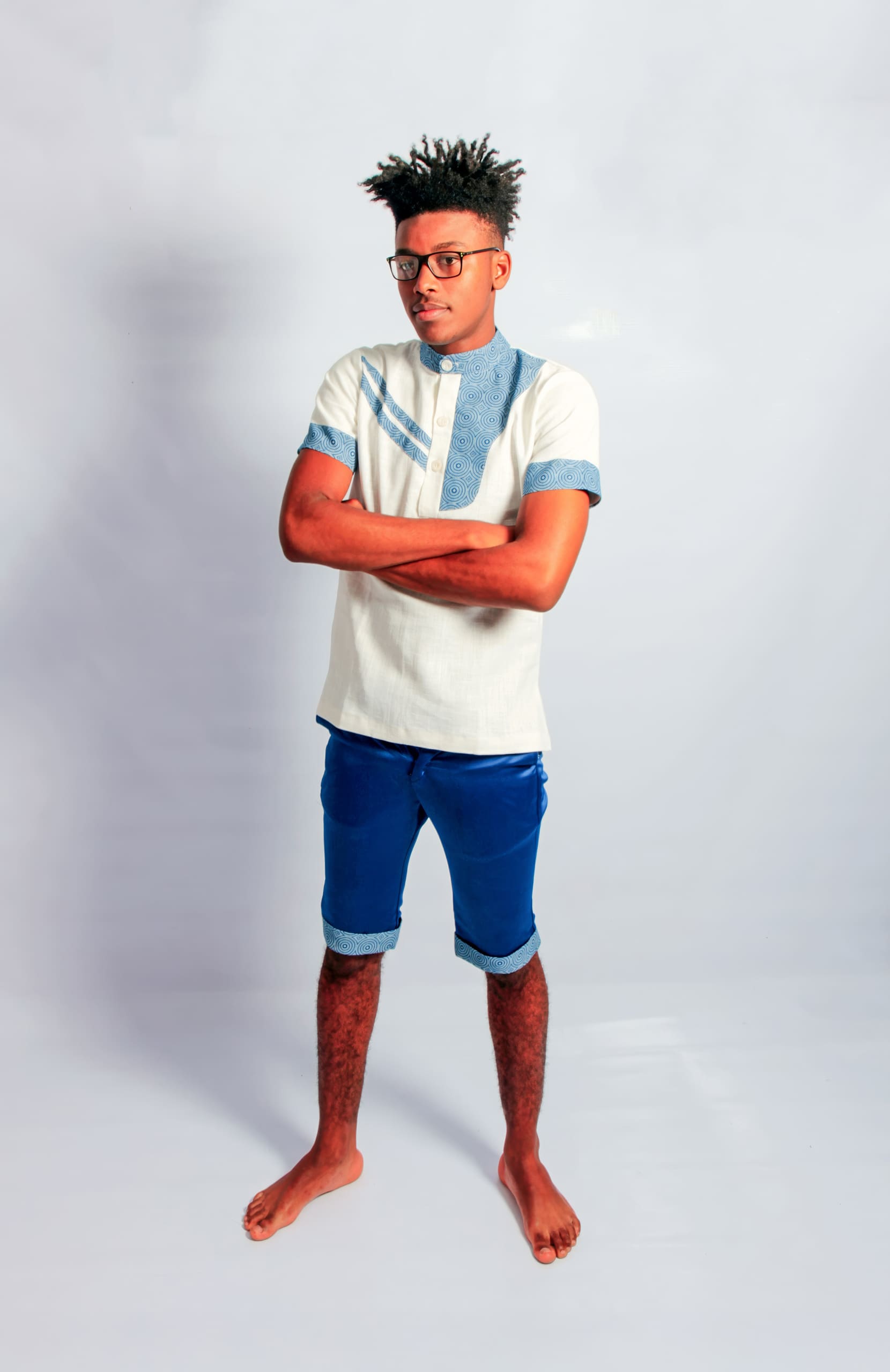 A young man with short, spiked hair and glasses stands barefoot against a plain white background. He wears a white shirt with blue trim and blue knee-length shorts, and has his arms folded, looking slightly to the side with a neutral expression.