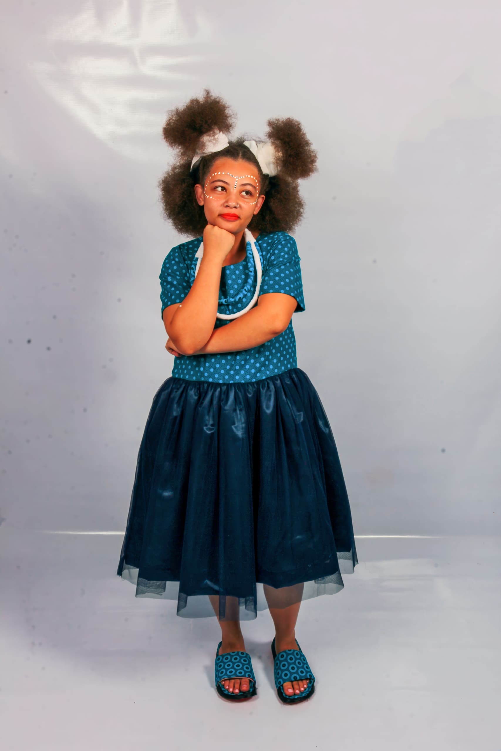 A young girl with large curly puffs and white bows stands thoughtfully with a hand on her chin. She wears round glasses, a teal spotty top, a dark blue tulle skirt, blue sandals, and a beaded necklace against a plain light background.