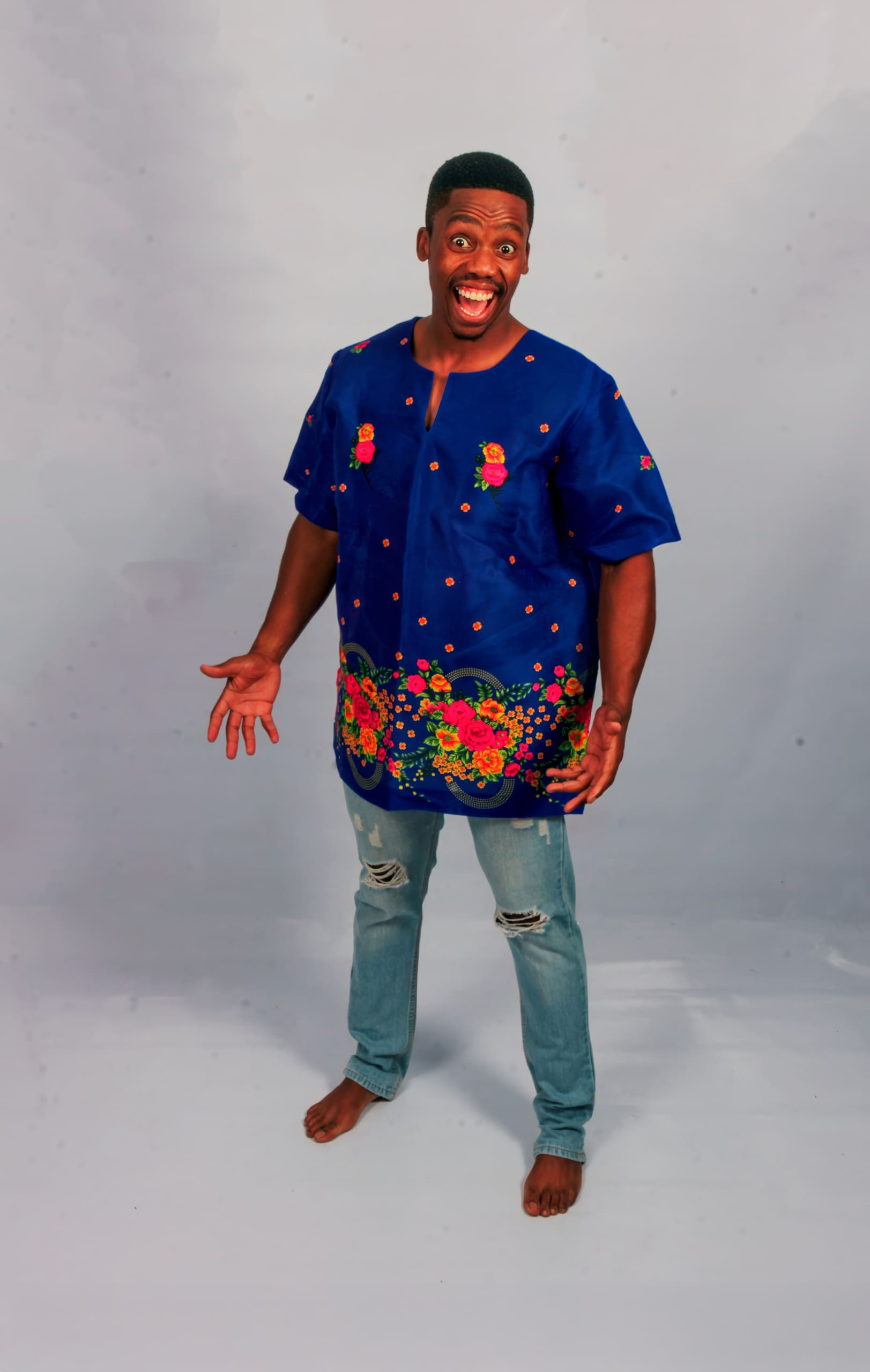 A smiling man stands barefoot on a light grey background, wearing a bright blue shirt with colourful floral patterns and ripped light blue jeans. His arms are open and expressive, and he looks cheerful and energetic.