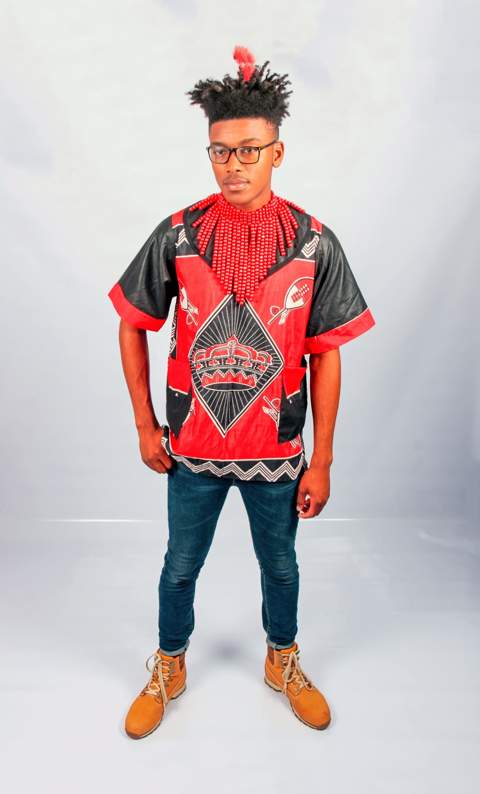 A young man stands confidently against a plain white background, wearing a red and black patterned shirt, blue jeans, tan boots, red beaded necklaces, and glasses. His hair is styled upwards with a red ornament.