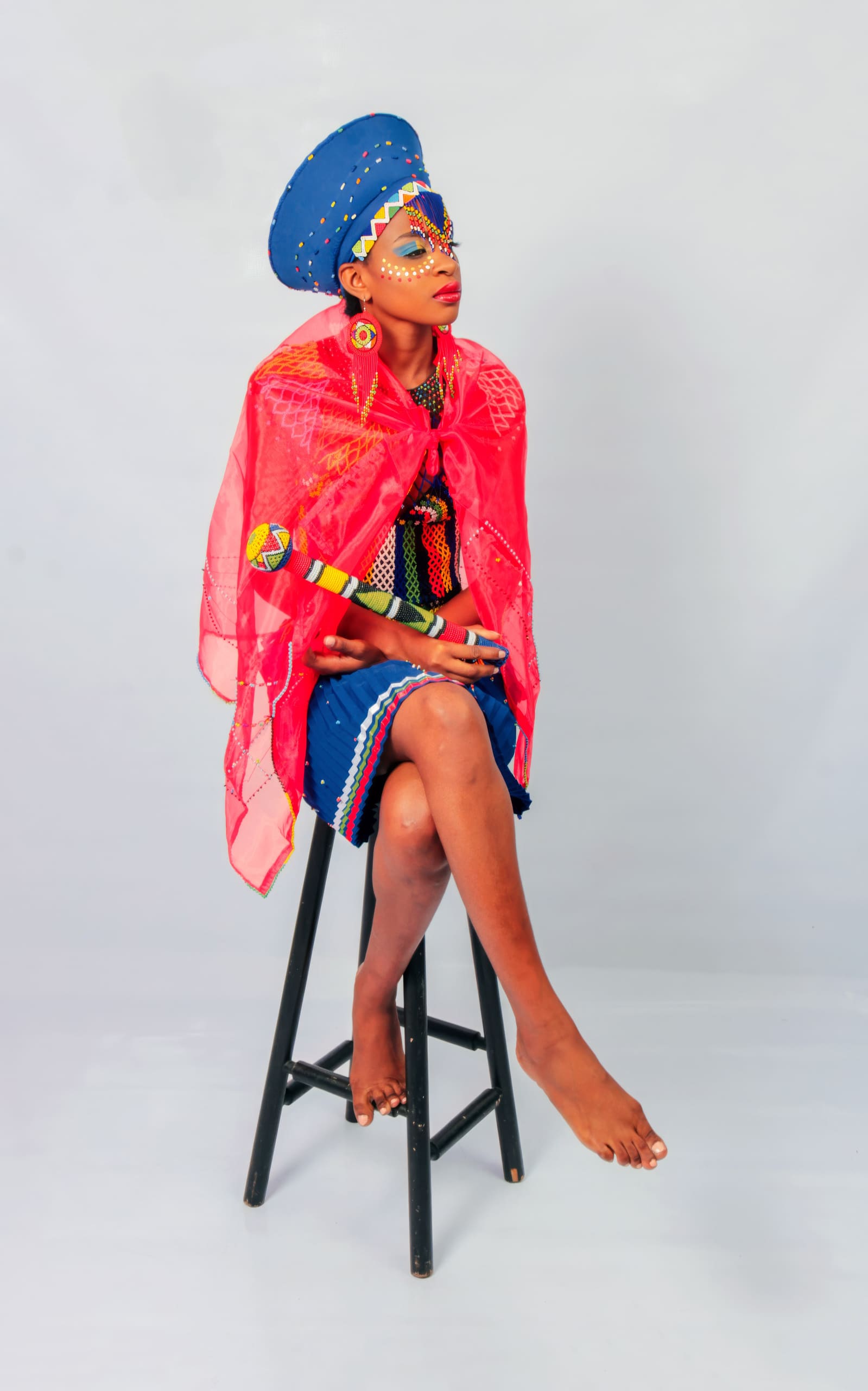 A person in bright traditional attire sits barefoot on a stool. They wear a blue, beaded headpiece, red sheer shawl, and colourful dress. Their face is painted with white patterns, and they look up and to the side against a plain backdrop.
