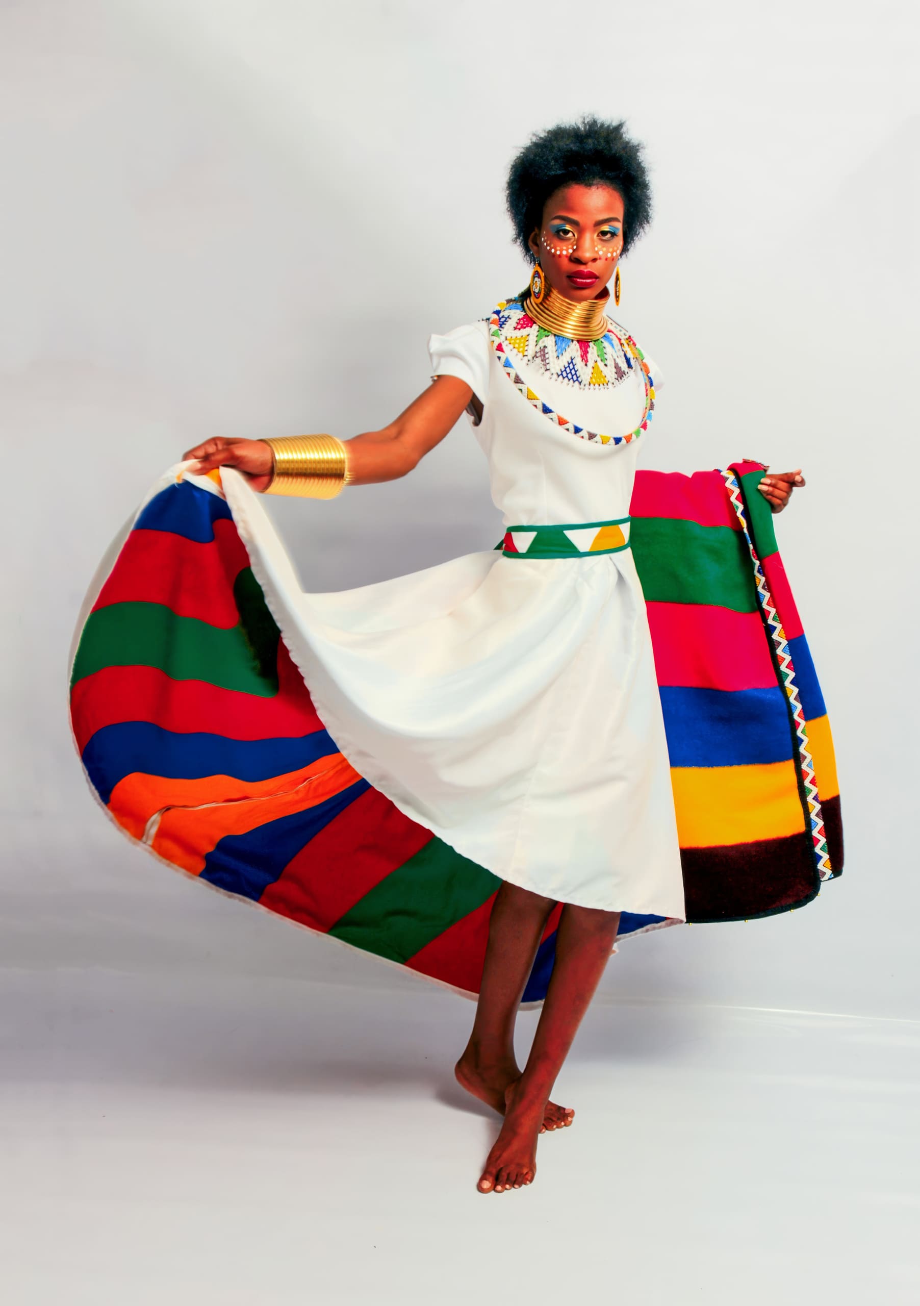 A barefoot woman poses in a white dress with colourful stripes on the skirt. She wears bold beaded jewellery, gold bangles, and traditional face paint. Her hair is natural, and she holds out her skirt to show off the vibrant design against a plain background.