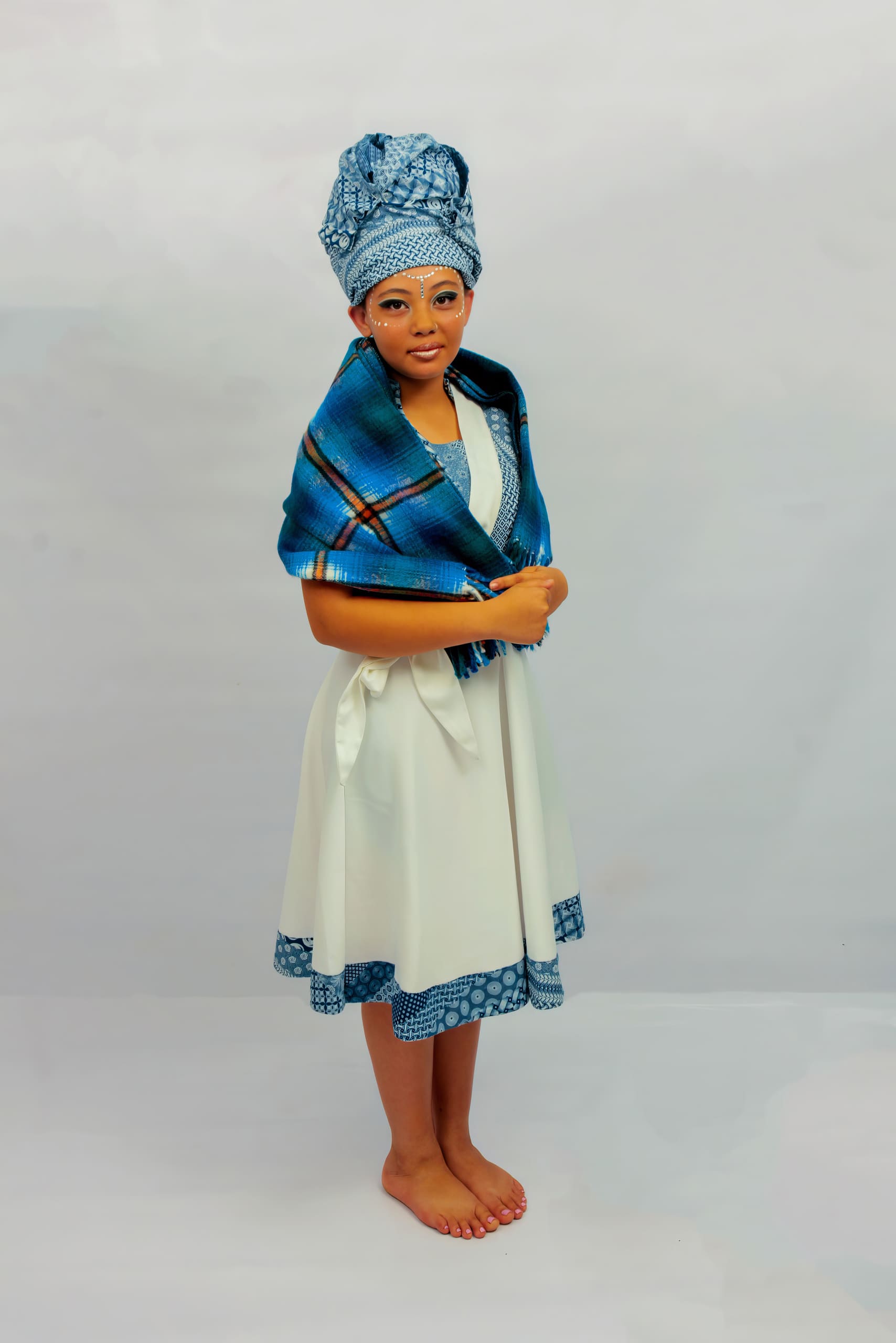 A young woman stands barefoot on a plain background wearing a white dress with blue patterned trim, a matching blue patterned headwrap, and a blue plaid shawl draped over her shoulders. She faces forwards with her hands lightly holding the shawl.