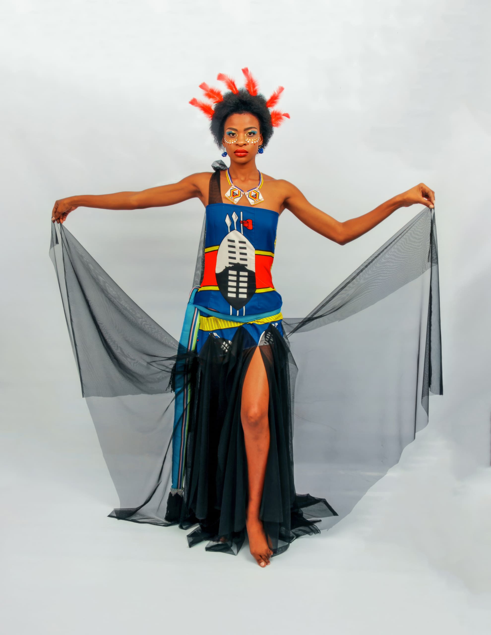 A woman stands with arms outstretched, holding sheer black fabric. She wears a colourful dress with bold patterns and a tall headdress decorated with red accents. Her make-up is bright and artistic, and she poses barefoot against a plain white background.