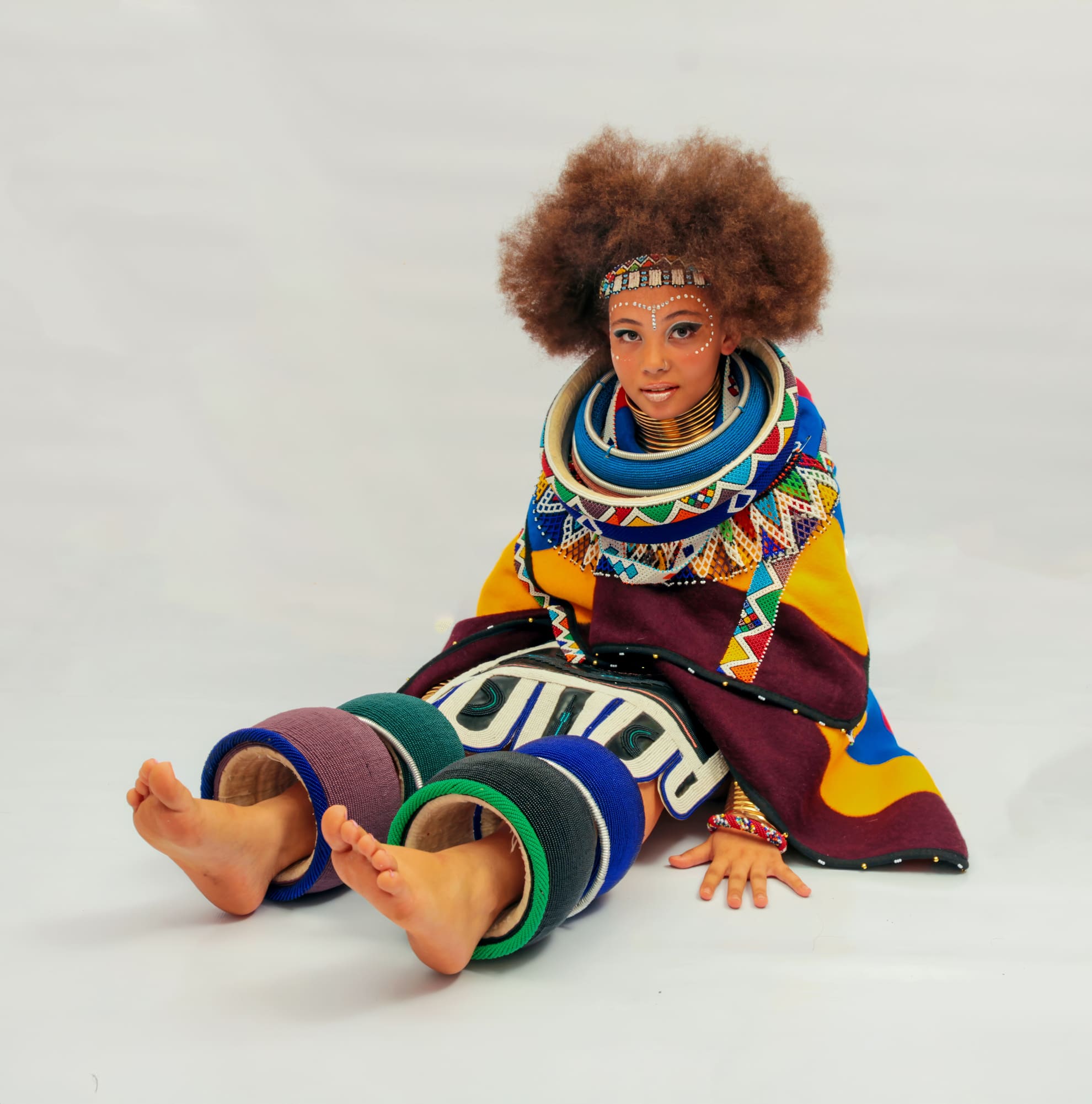 A person with curly hair sits on the floor against a white background wearing traditional, colourful Ndebele attire with beadwork, neck rings, arm rings, leg rings, and a patterned blanket. Their face has detailed painted designs, and they look towards the camera.