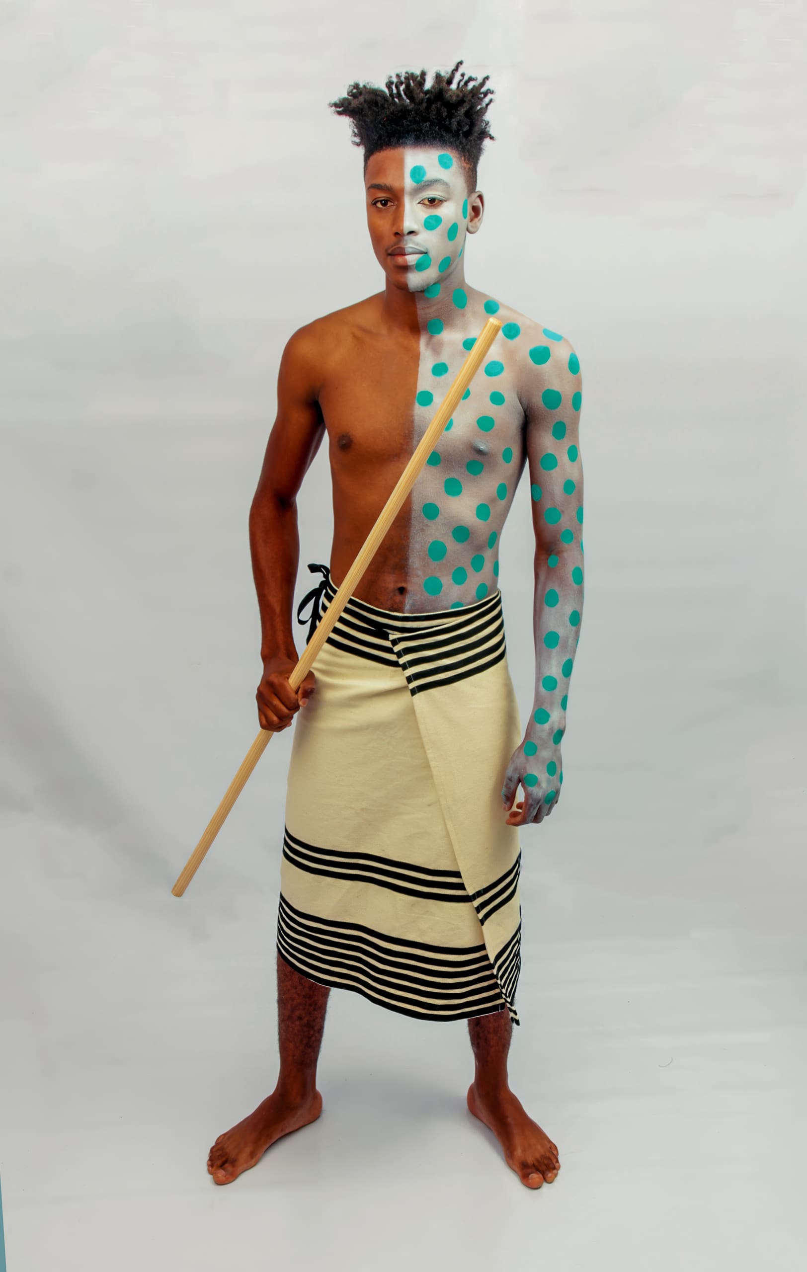 A barefoot man stands against a plain white background, wearing a cream-coloured wrap with black stripes. His upper body and face are painted half blue and half white, with blue polka dots on the white side. He holds a wooden staff vertically in one hand.